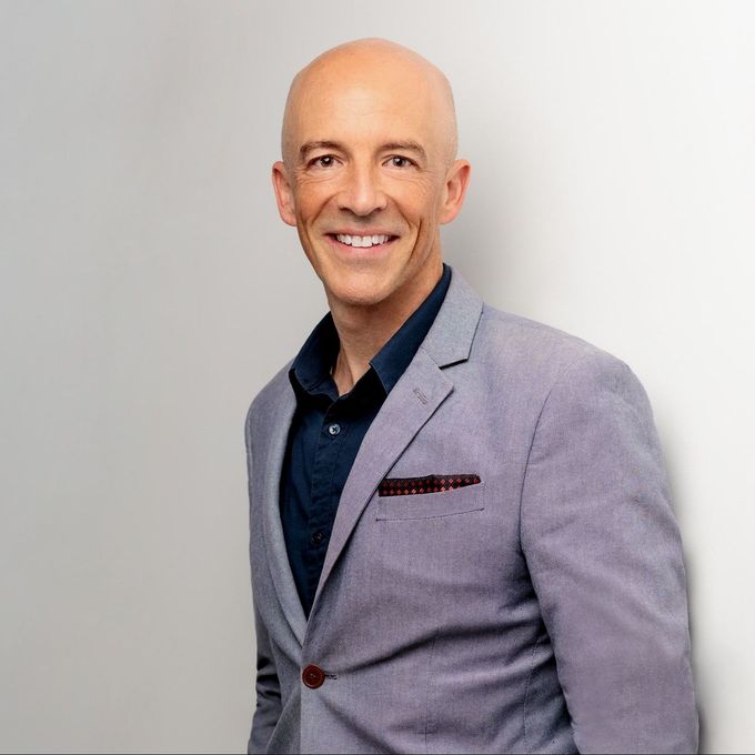 A bald man in a suit is smiling and standing in front of a white wall.