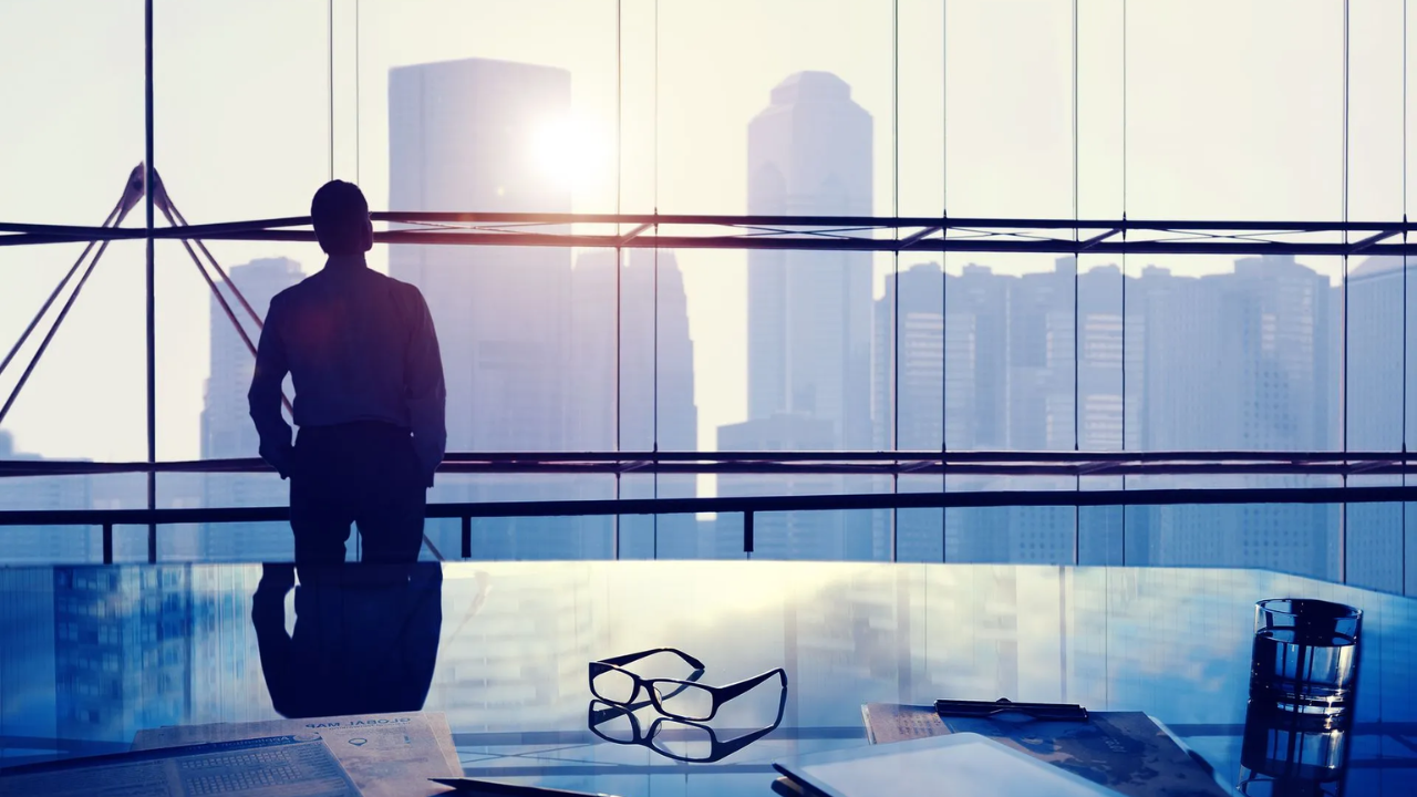 Silhouette of a person looking out a large window at a cityscape from inside a modern office.