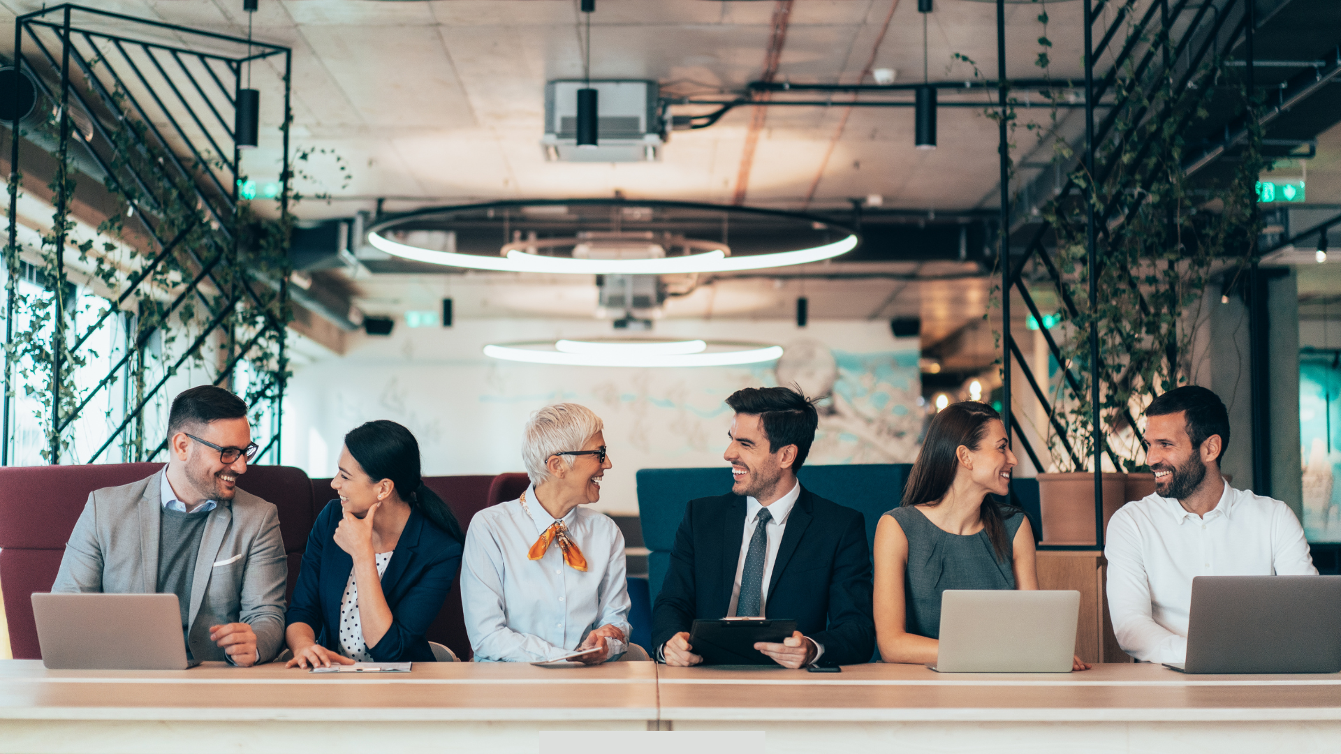 A diverse group of six professionals seated at a long table in a modern office, collaborating and talking while working.