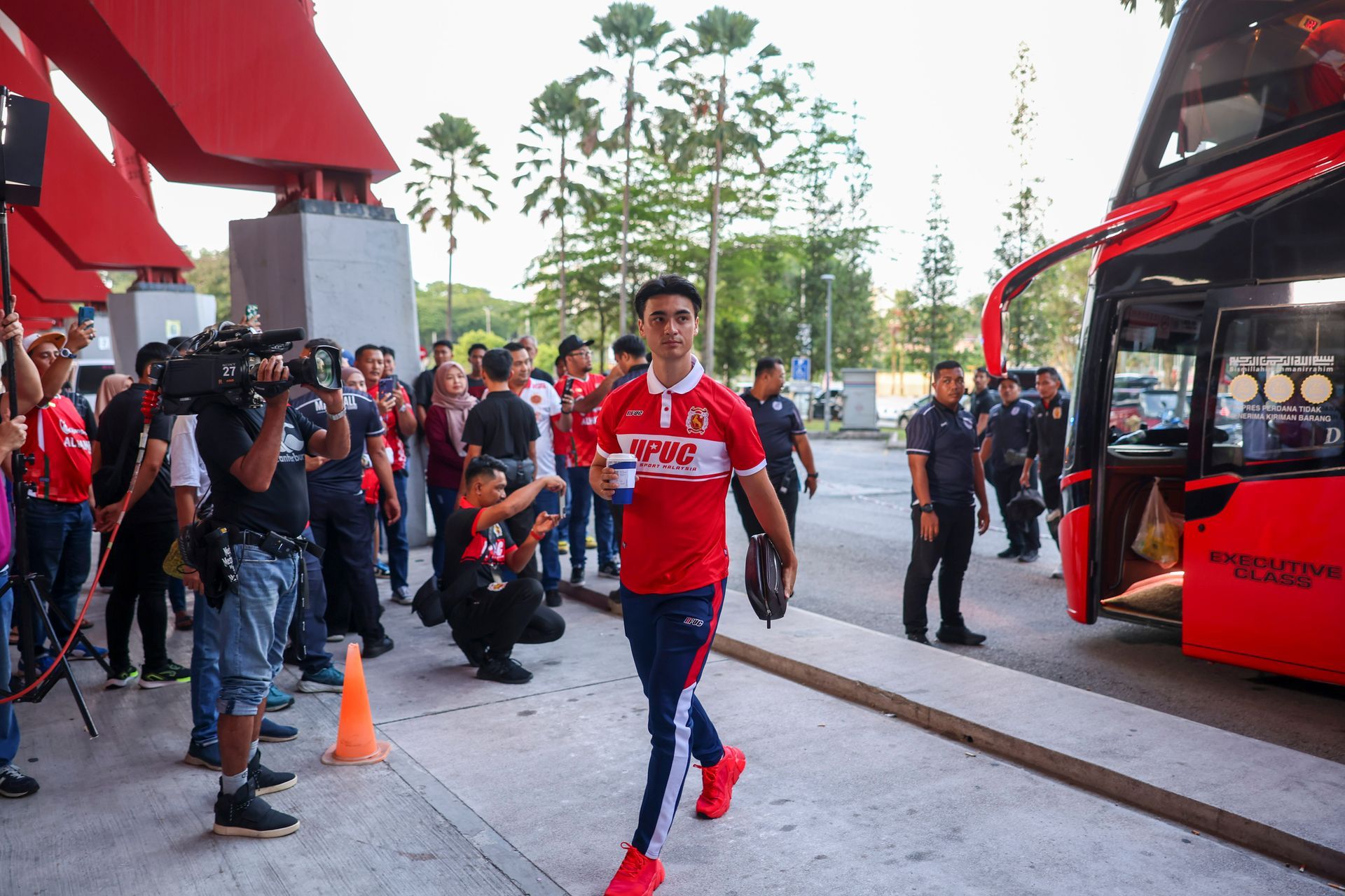 A soccer player in a red uniform walks past a red bus, surrounded by media and fans.
