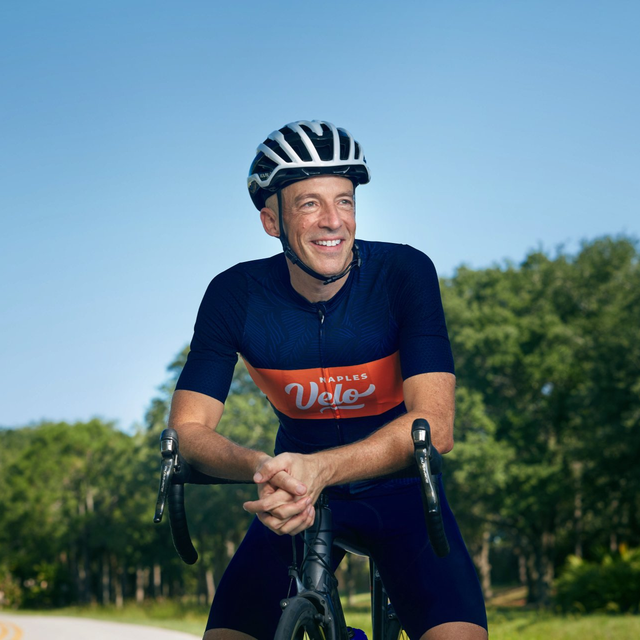 Michael Ceely, high performance coach smiling, resting on a road bike outdoors on a sunny day.