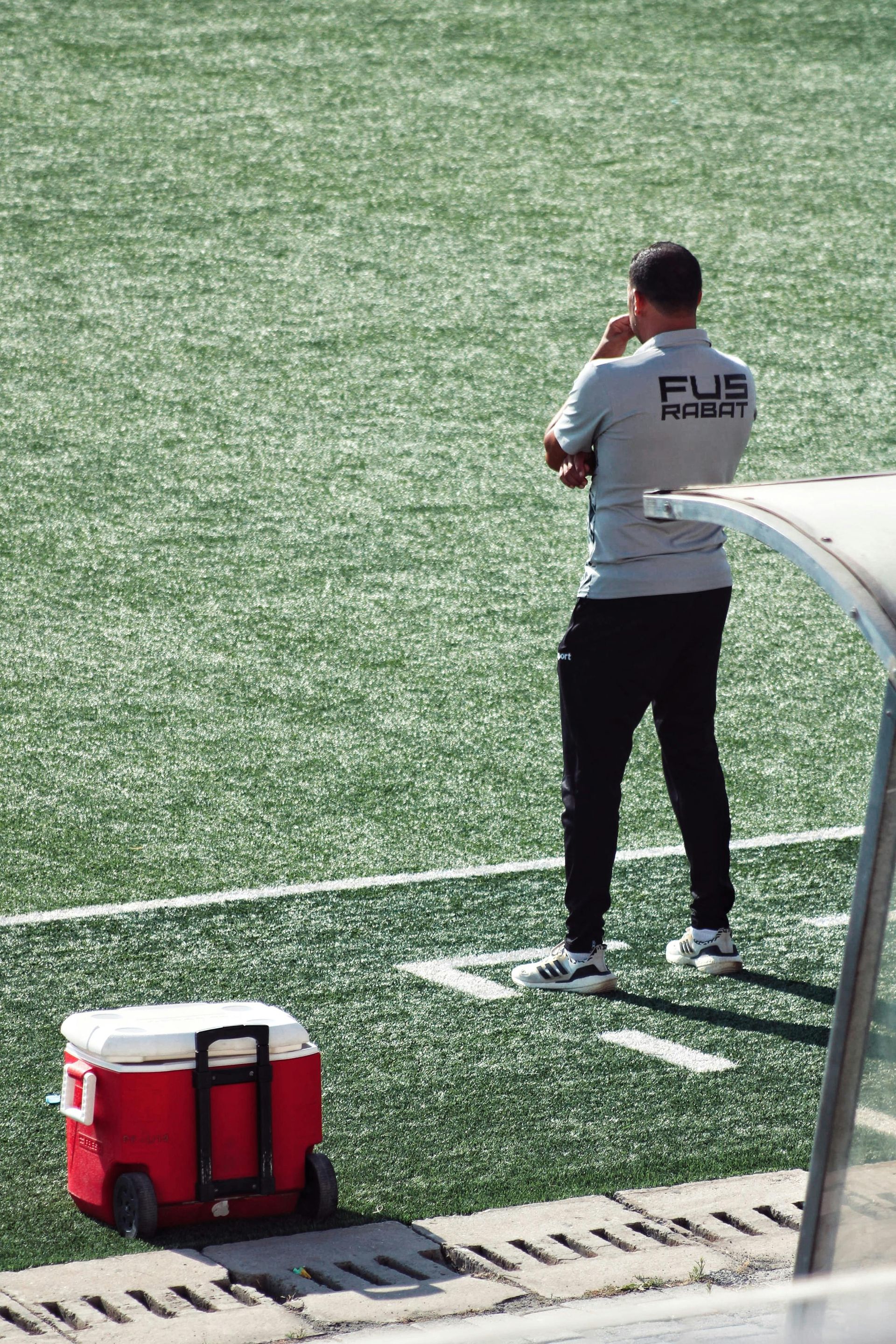 Coach on a soccer field, looking on with a cooler nearby.