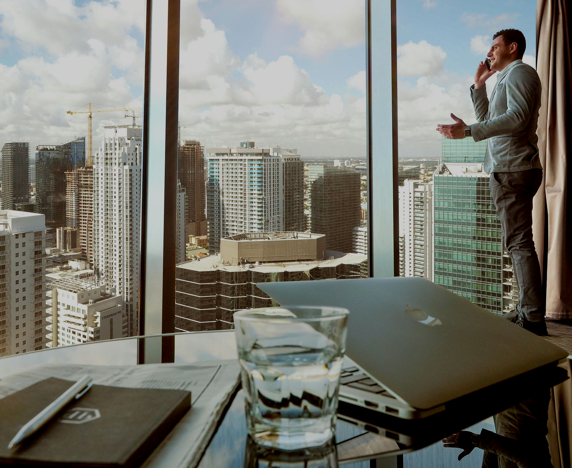 Businessman talking on the phone, looking out at a cityscape through a window behind a desk with laptop and a glass of water.