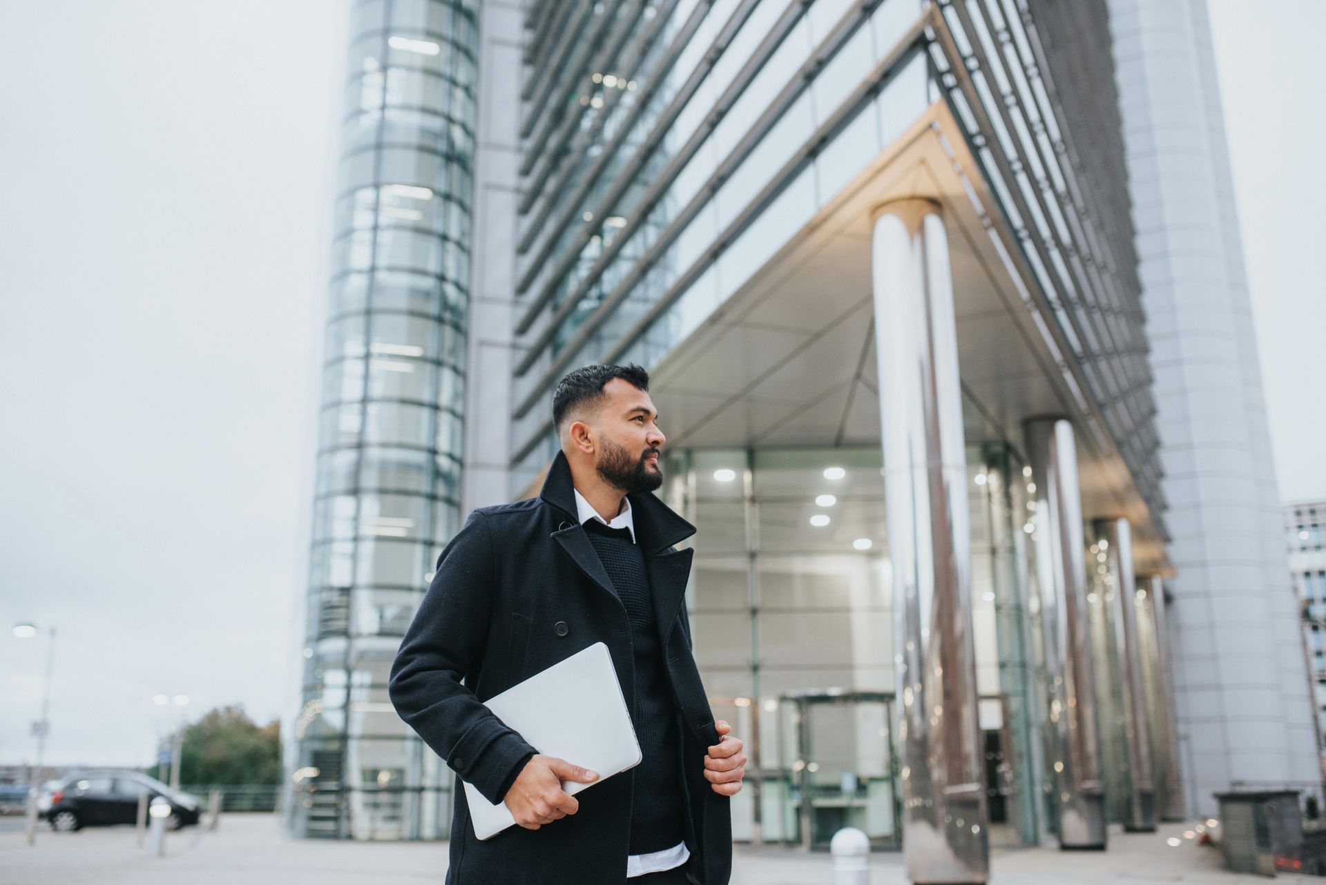 Man with laptop, standing outside modern glass building, looking away.