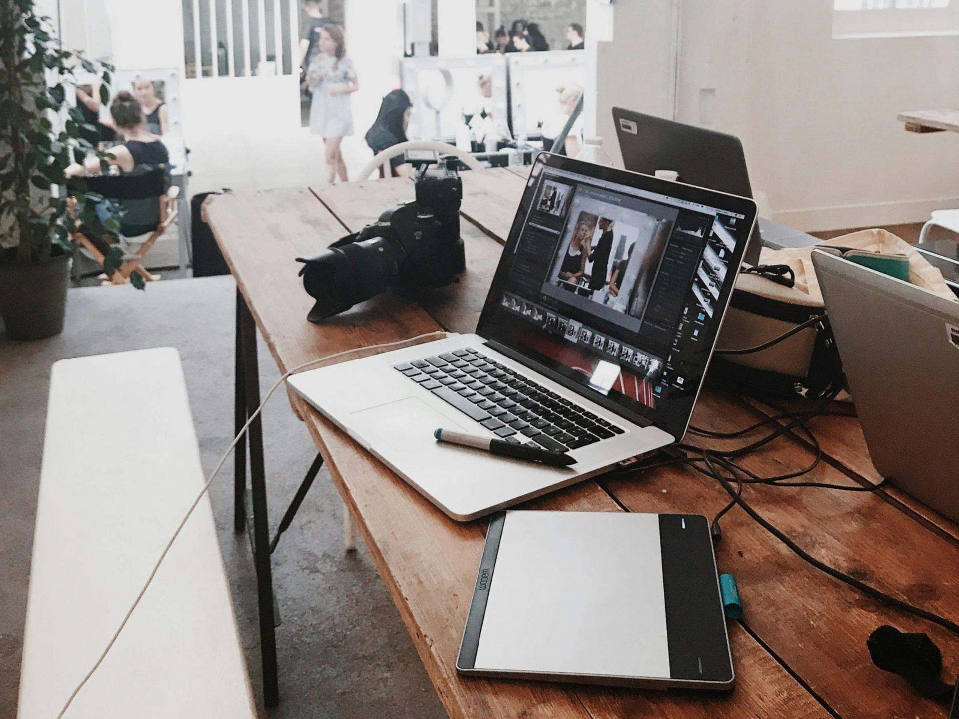 Laptop and camera on wooden table in a brightly lit studio. A person stands in the background.