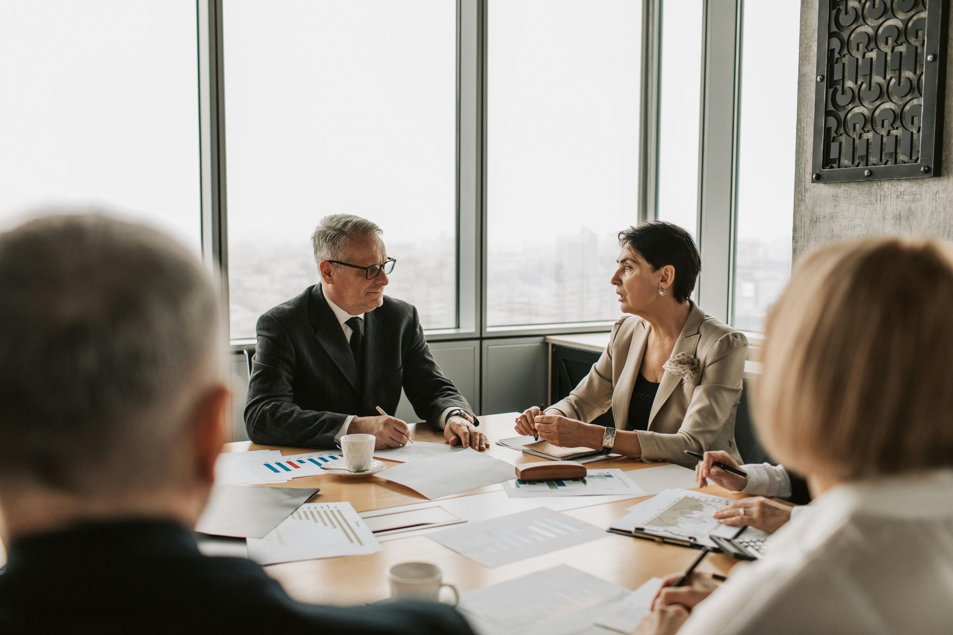 Business meeting in a modern office. People sit around a table, discussing documents, with a cityscape visible.