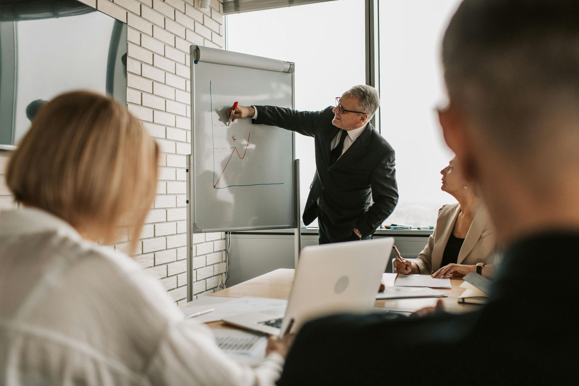 A person in a suit points to a whiteboard while speaking to colleagues in a bright office meeting.