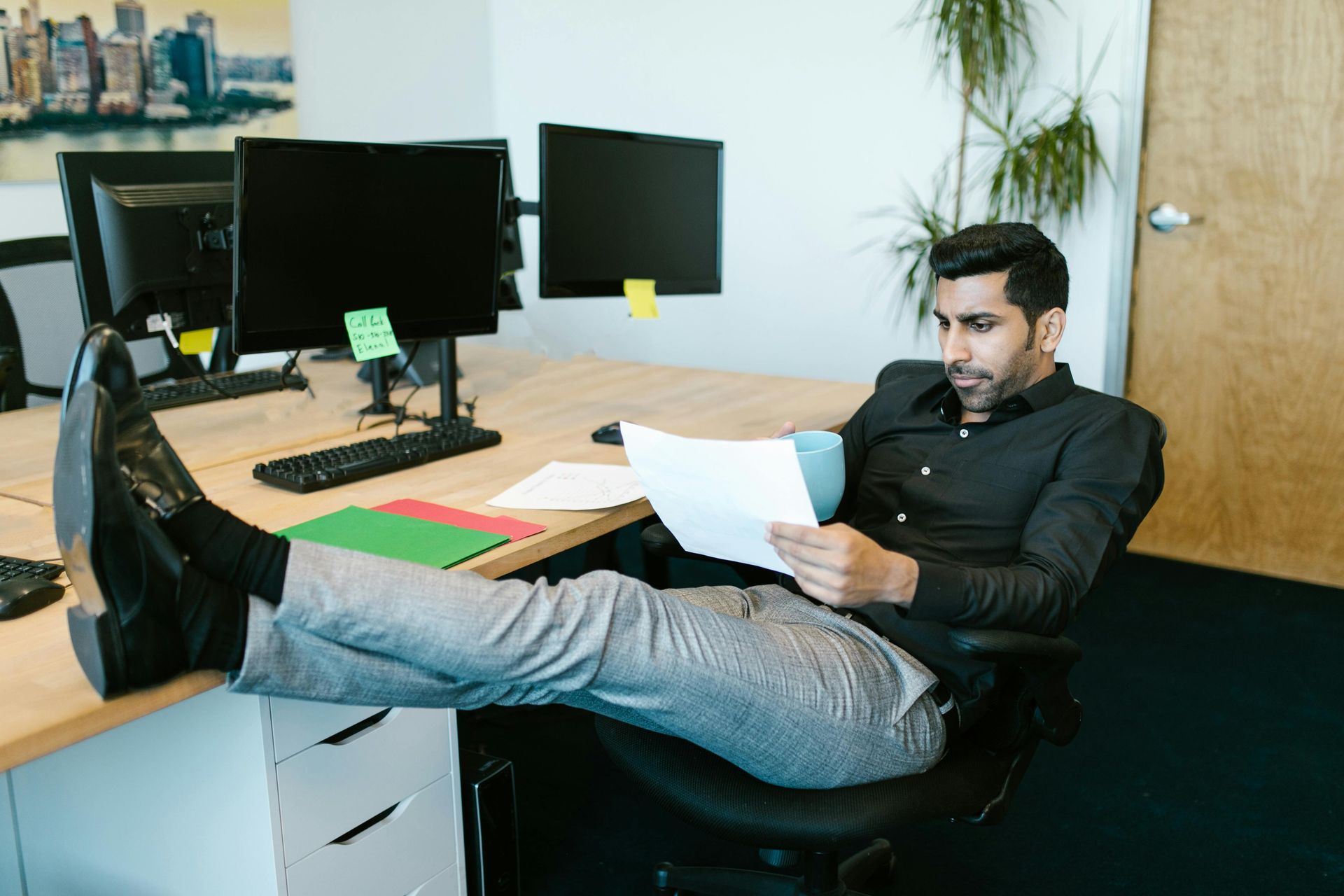 Man sitting at desk, feet up, reading papers. Office setting with computer monitors and papers.