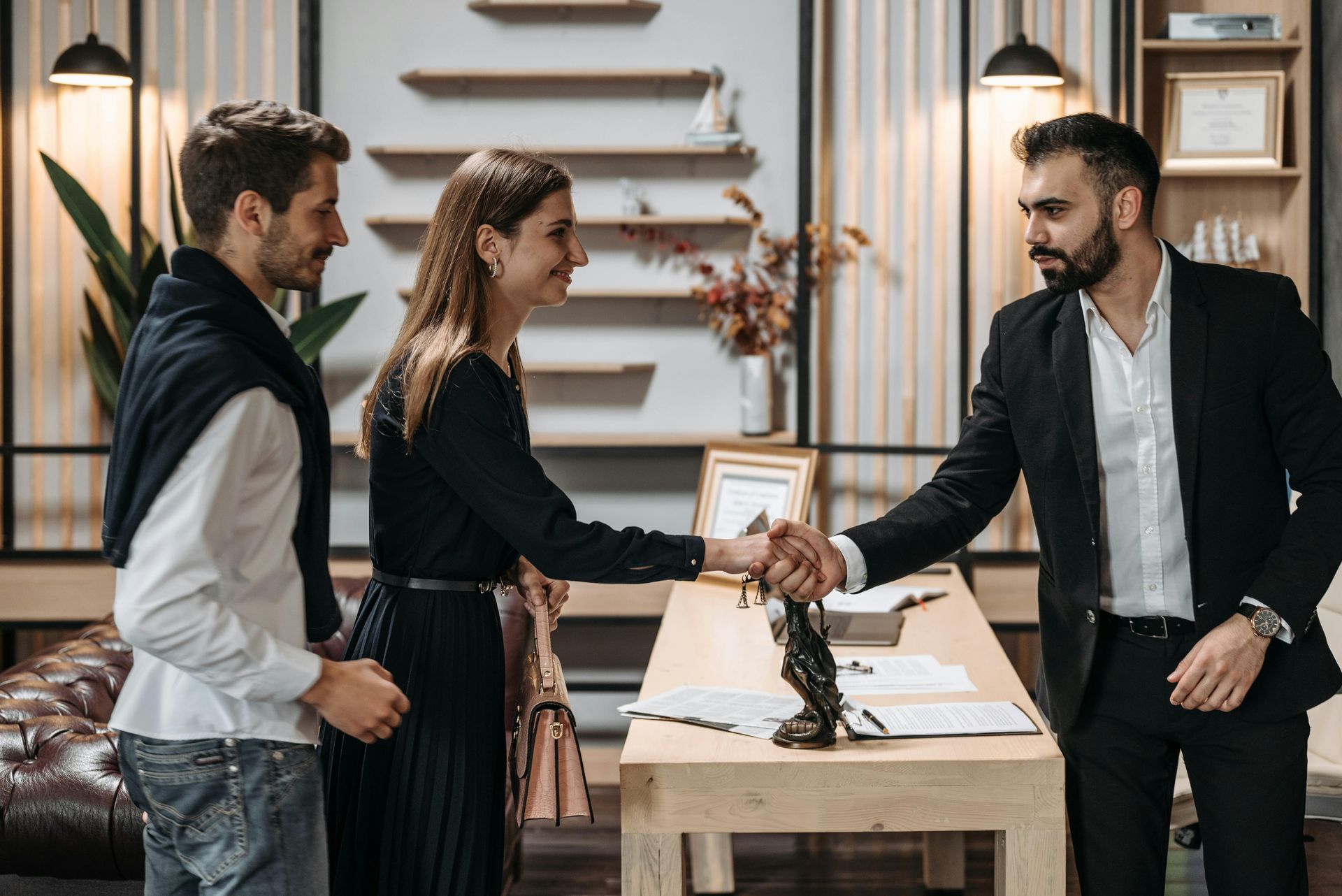 Man in suit shakes hands with woman, as another man looks on. Office setting.