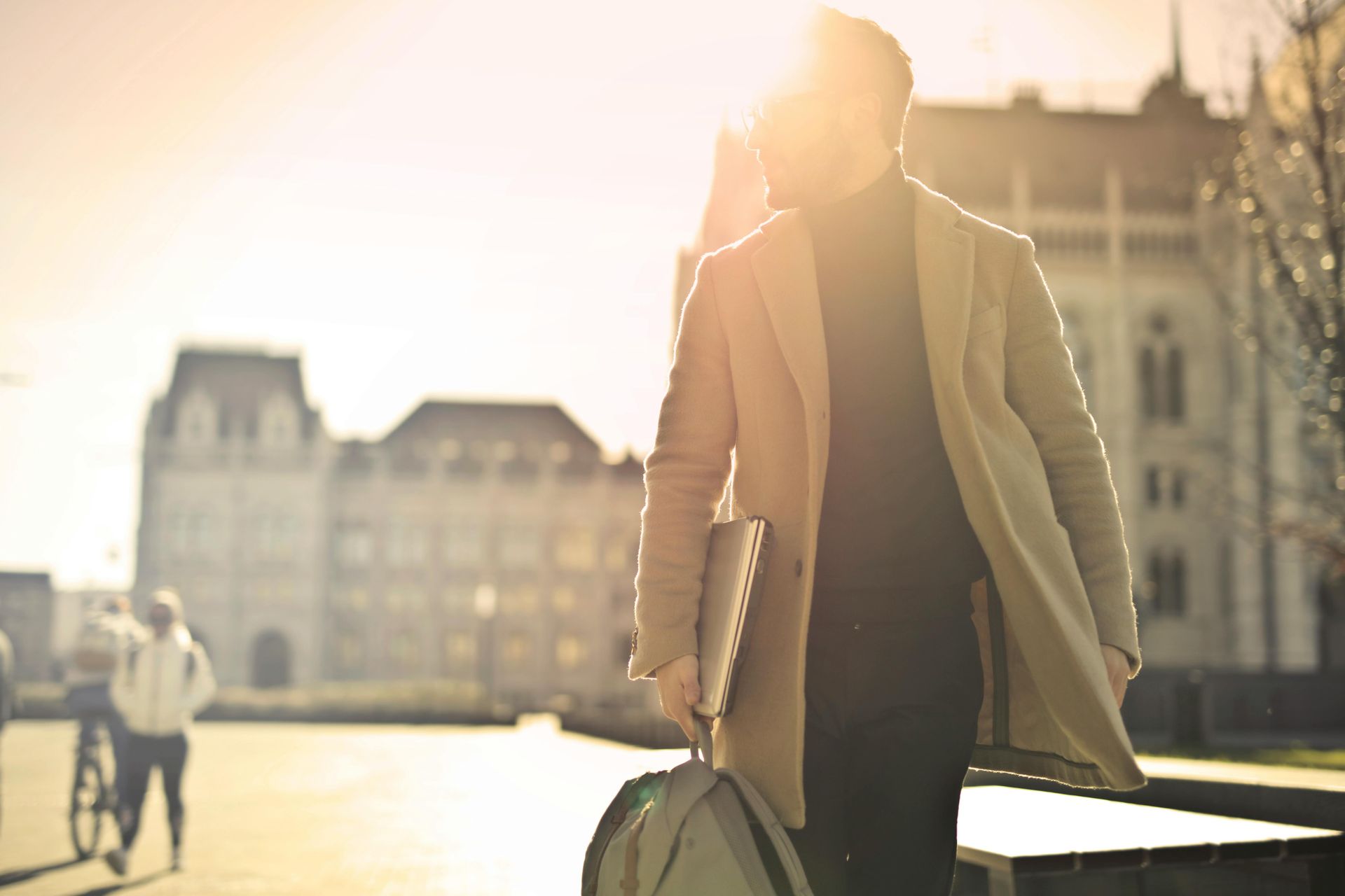 Man in coat walks outside, holding a laptop and bag. Sunny day, building in background.