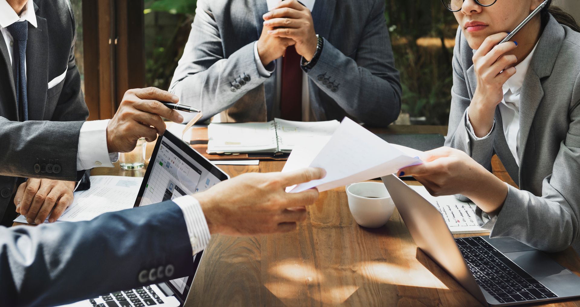 Business meeting around a wooden table. Hands passing documents and working on laptops.