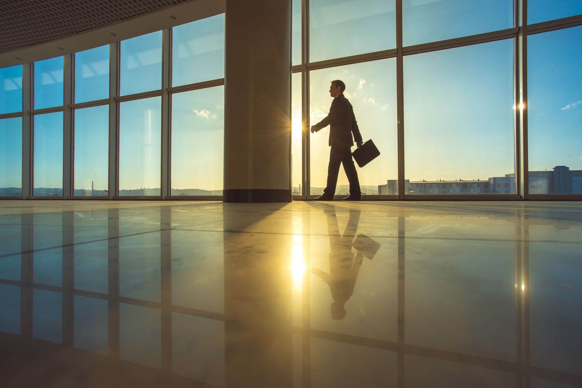 Businessperson walks in a bright office with large windows, holding a briefcase, with a reflection on the floor.