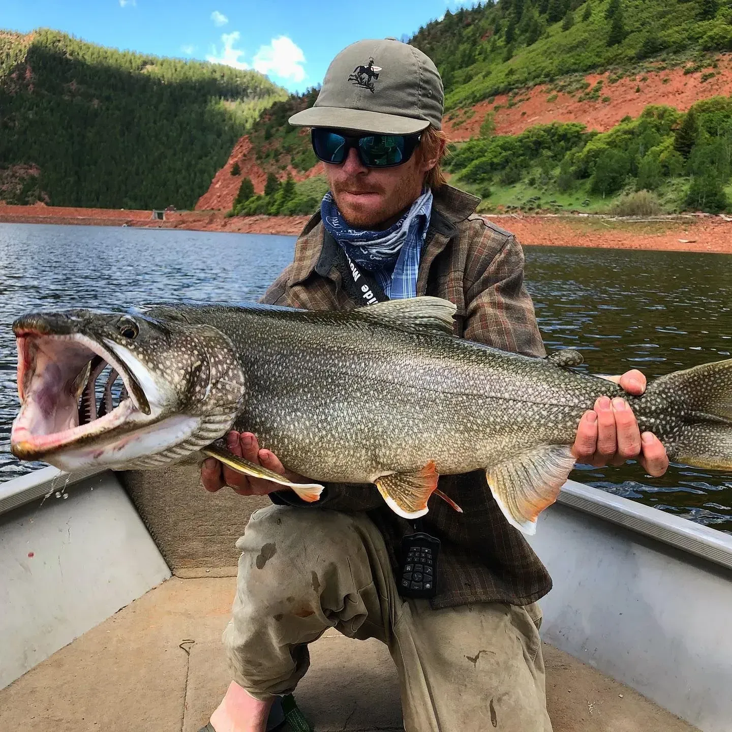 Man holding large fish in a boat, open mouth, lake and mountains in background.