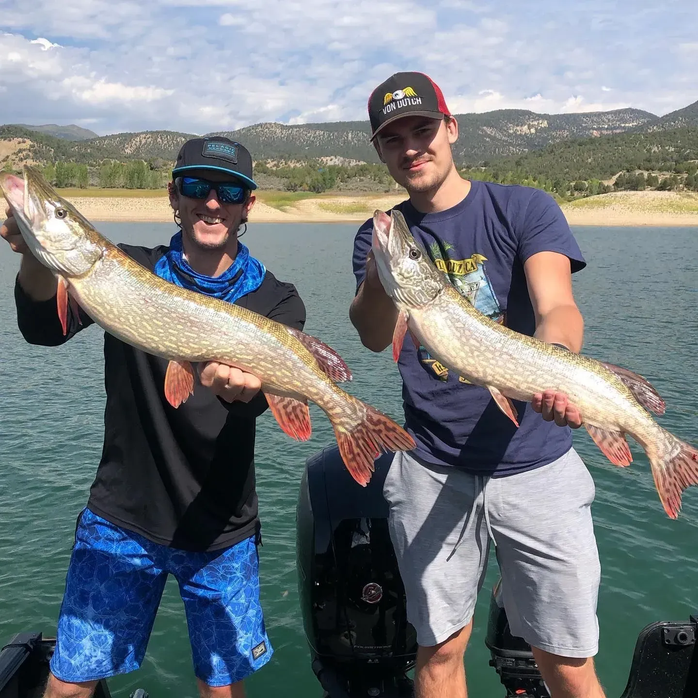 Two men on a boat hold up large fish they caught, with a lake and mountains in the background.
