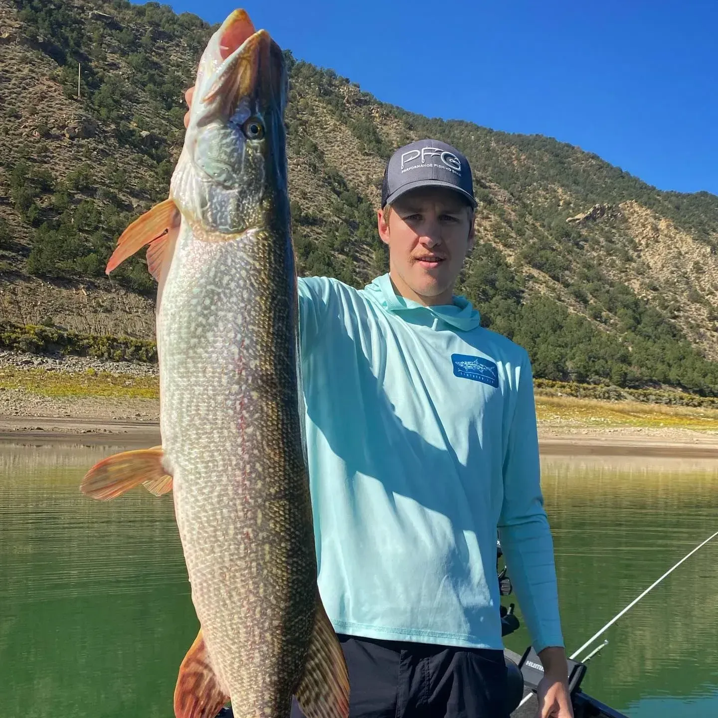 Man on boat holds up a large fish he caught, with mountains in the background.