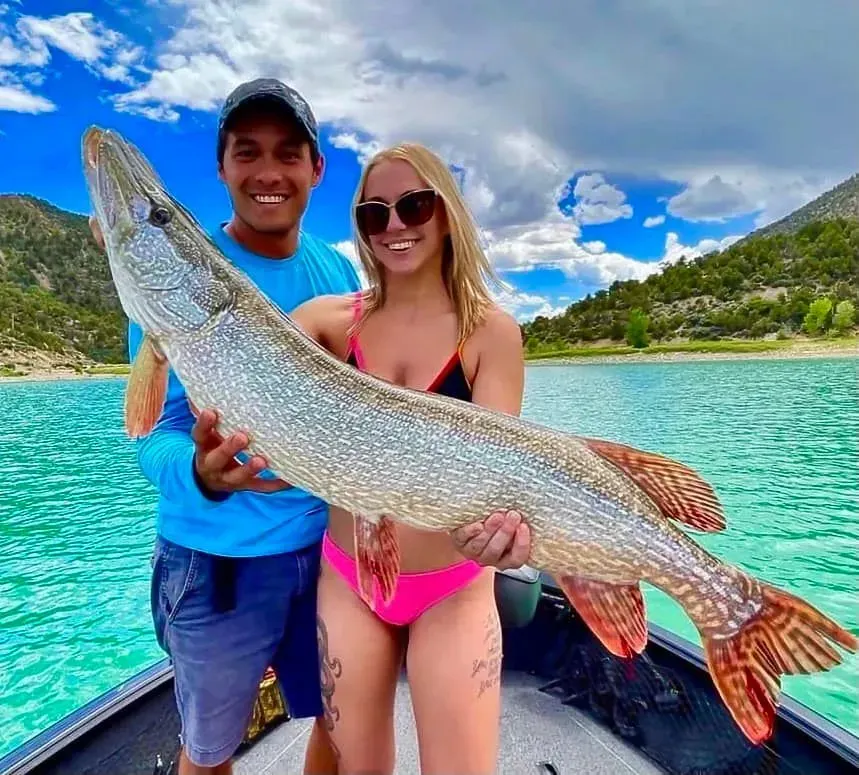Couple on a boat holding a large pike fish, with a lake and mountains in the background.