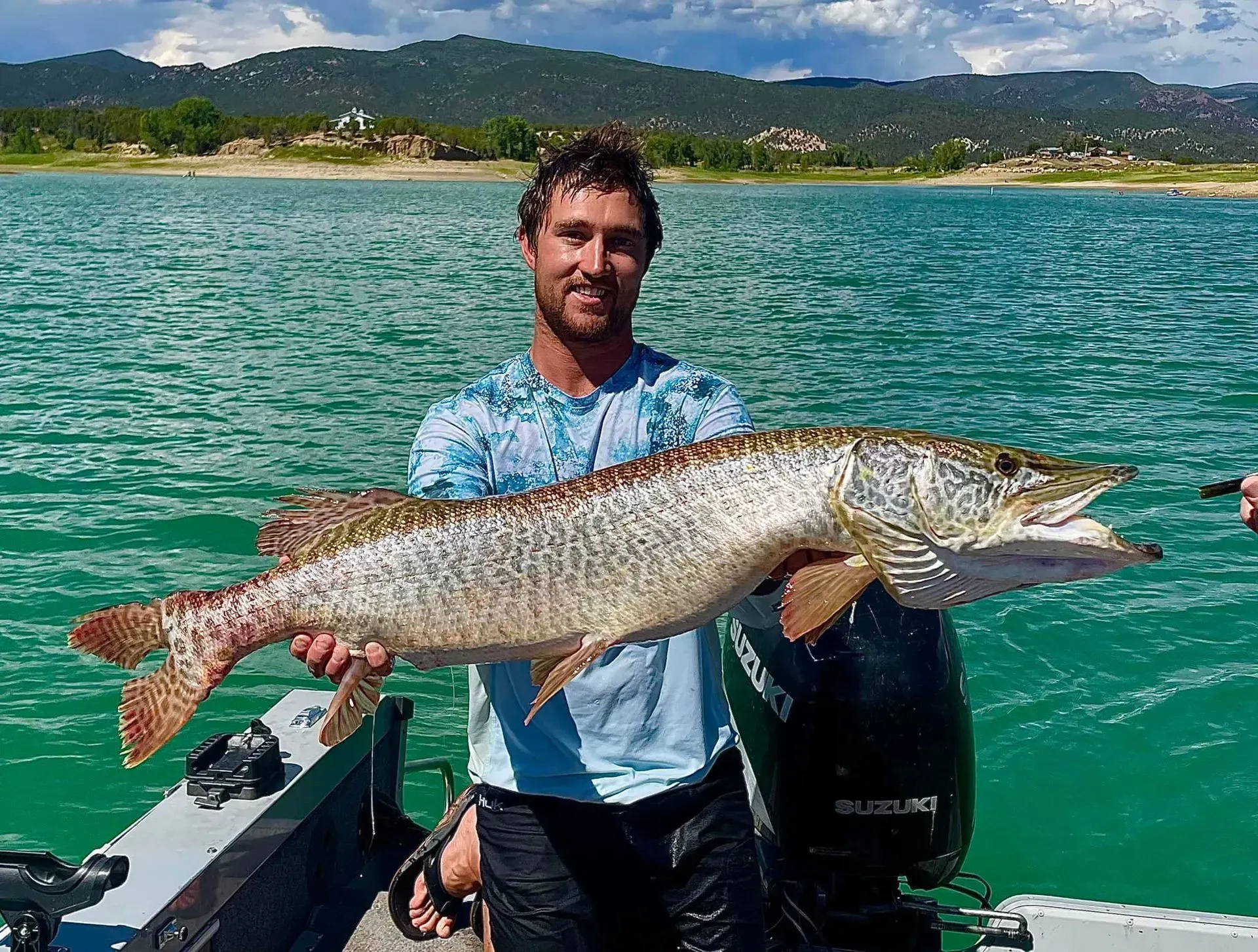 Man on a boat holding a large fish, possibly a muskie, with a lake and mountains in the background.