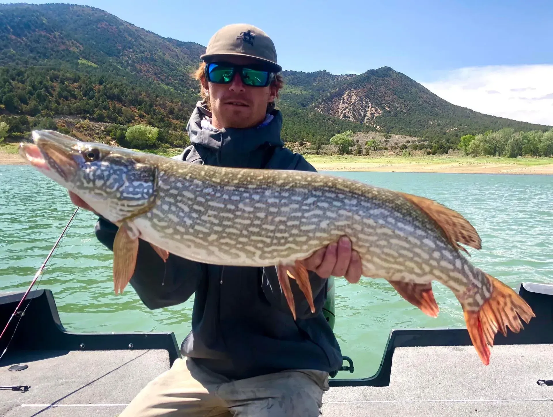 Man on boat holding large fish; mountains and lake in background.