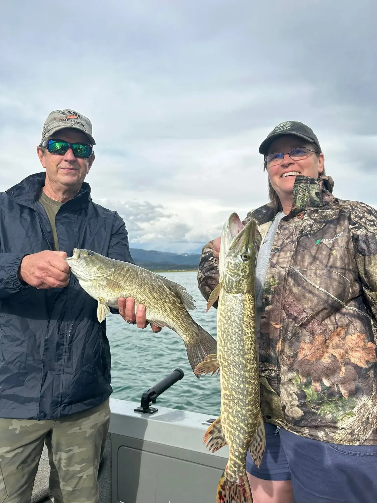 Two people on a boat holding fish they caught, with water and mountains in the background. Cloudy day.