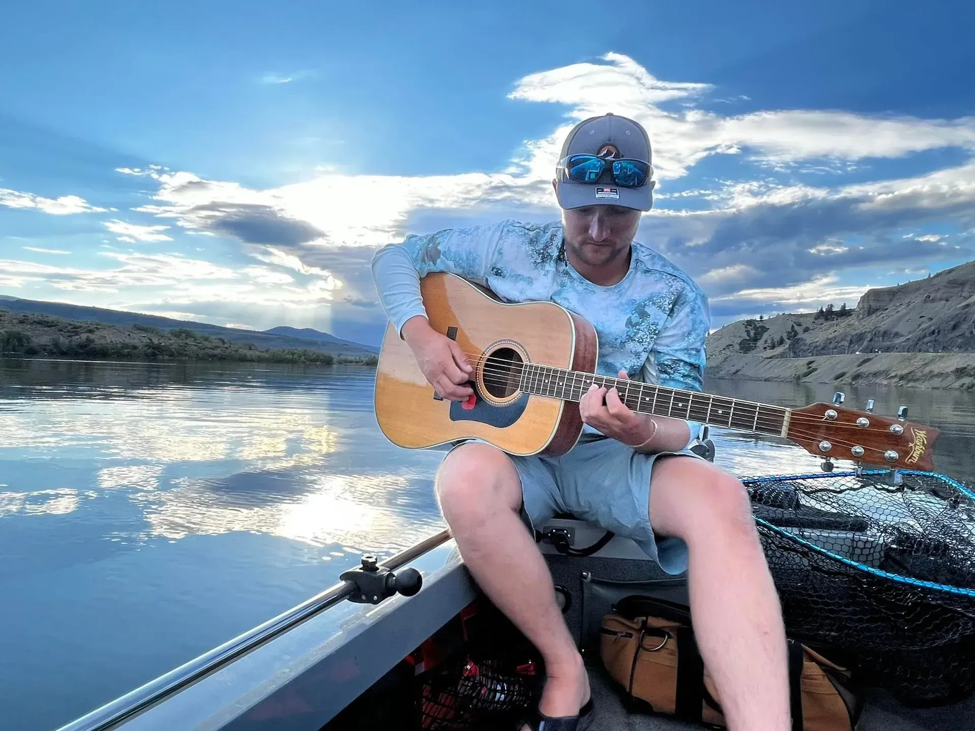 Man playing acoustic guitar in a boat on a lake under a bright, cloudy sky.