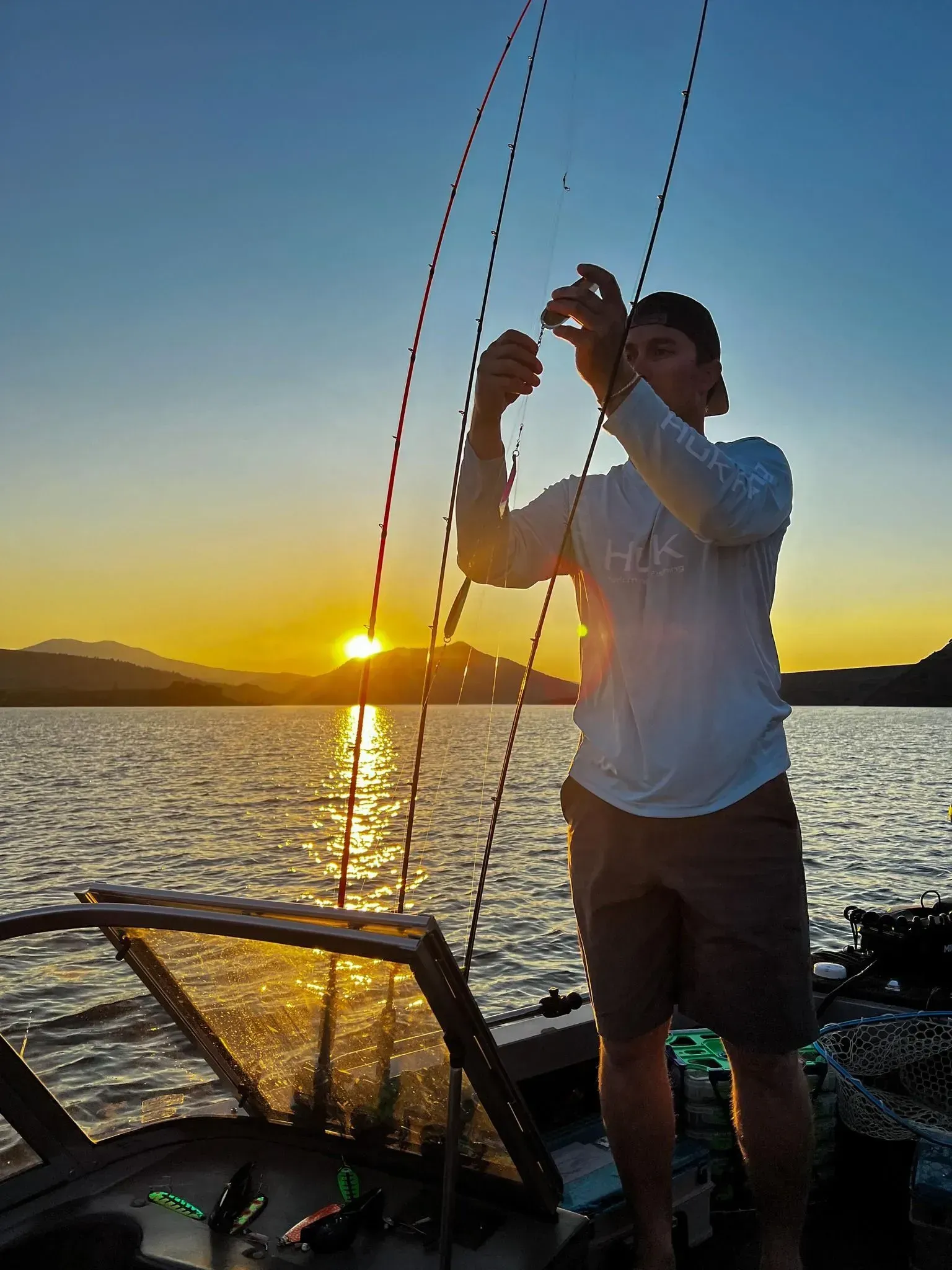 Man fishing at sunset, holding line. Lake, mountains, and orange sunlight in the background.