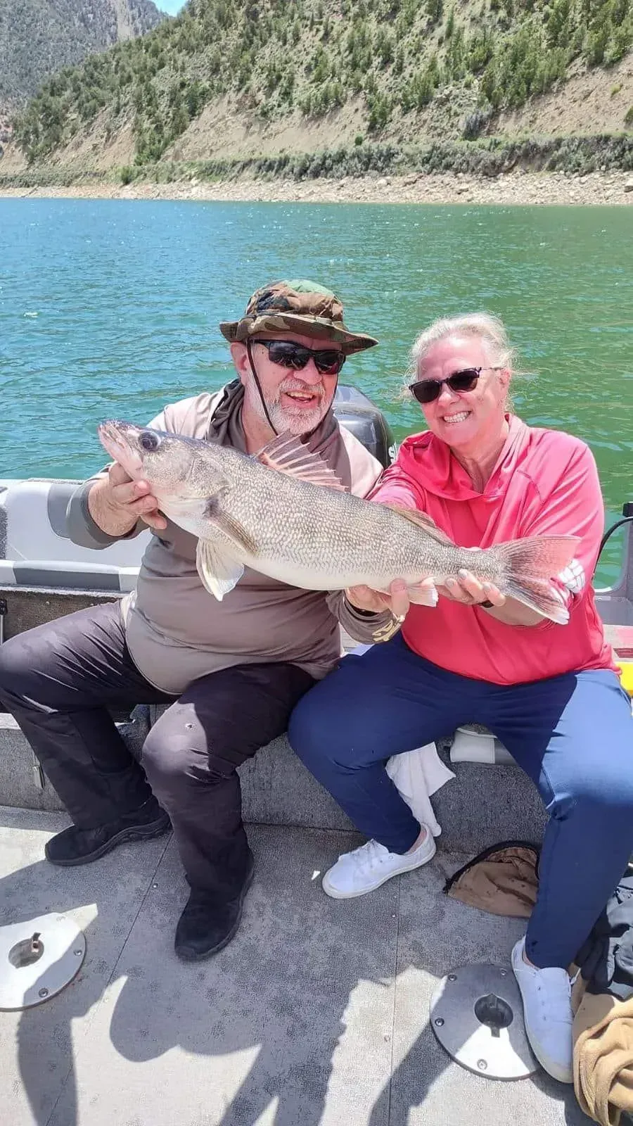 Two people on a boat holding a large fish near a body of water and mountains.