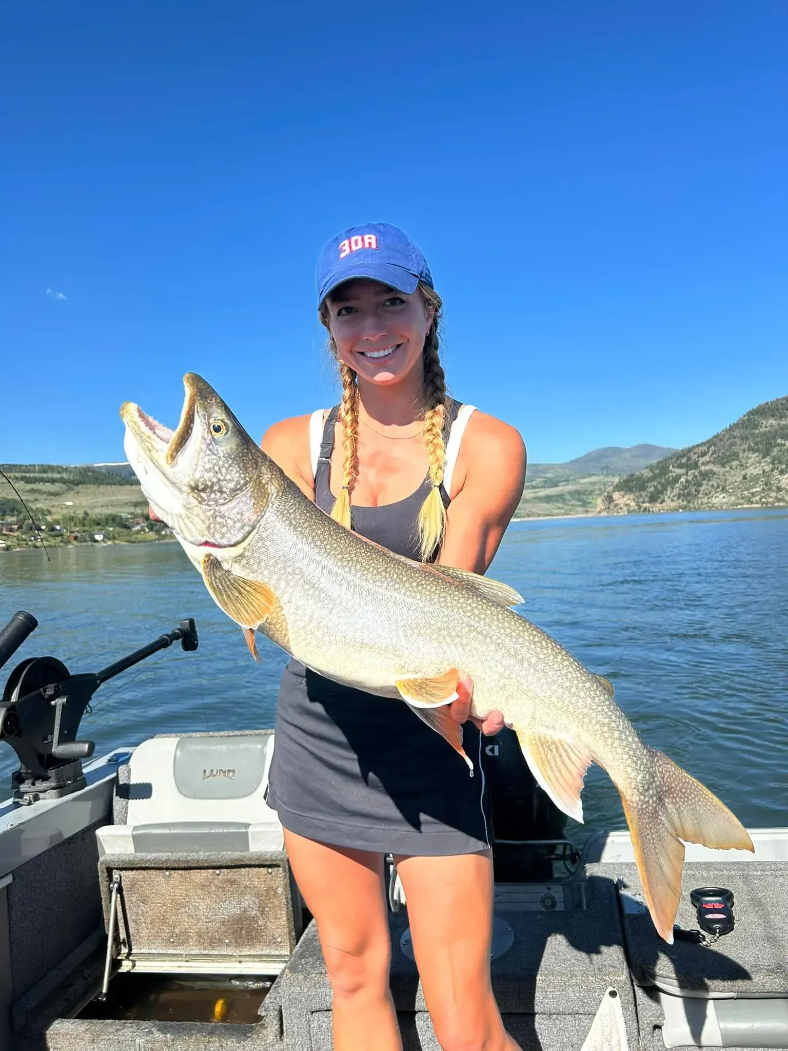 Woman holding large fish on a boat in sunny setting. Smiling, wearing hat and dress.