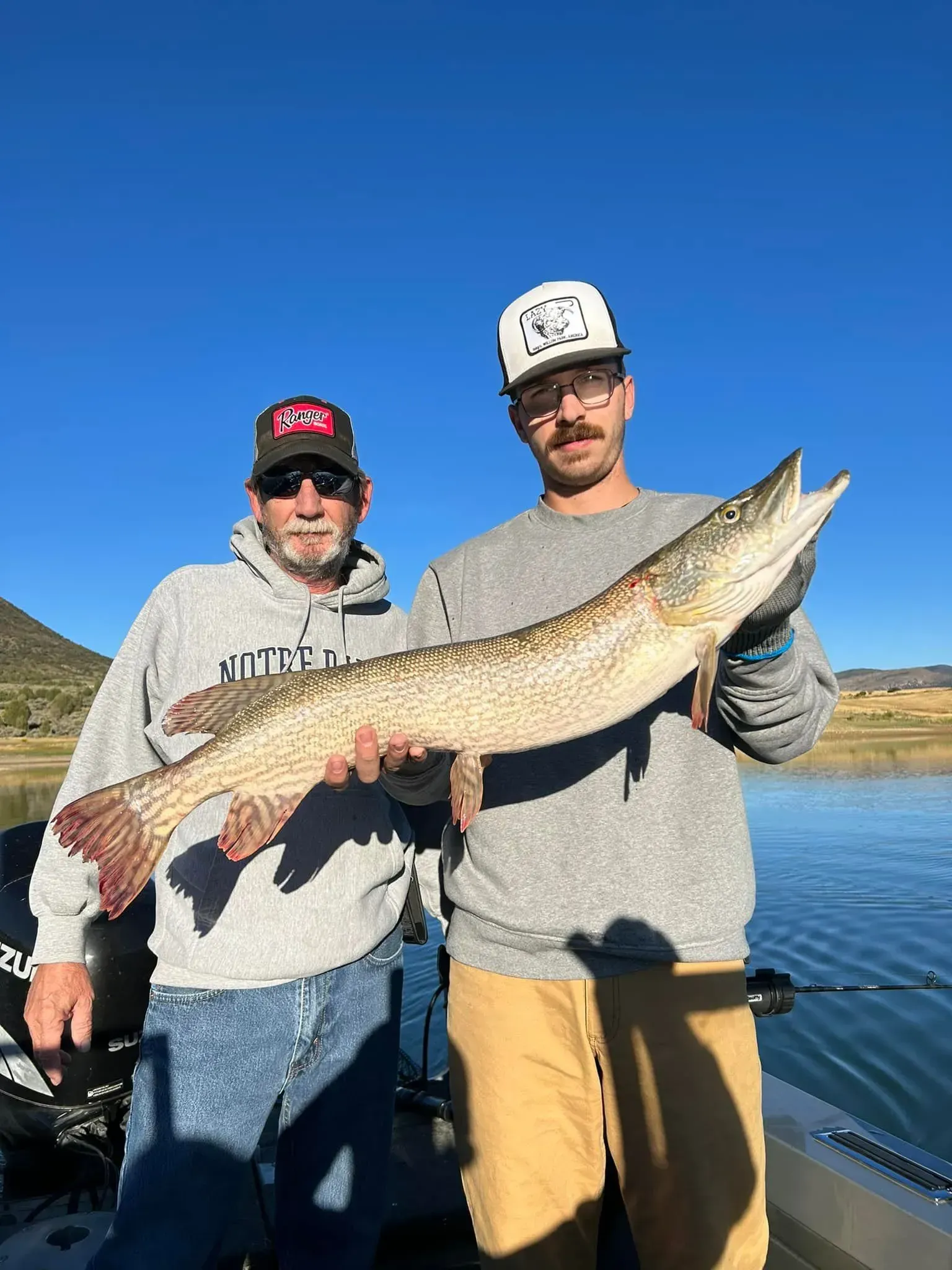 Two men on a boat hold up a large fish they caught. Blue sky and water in the background.
