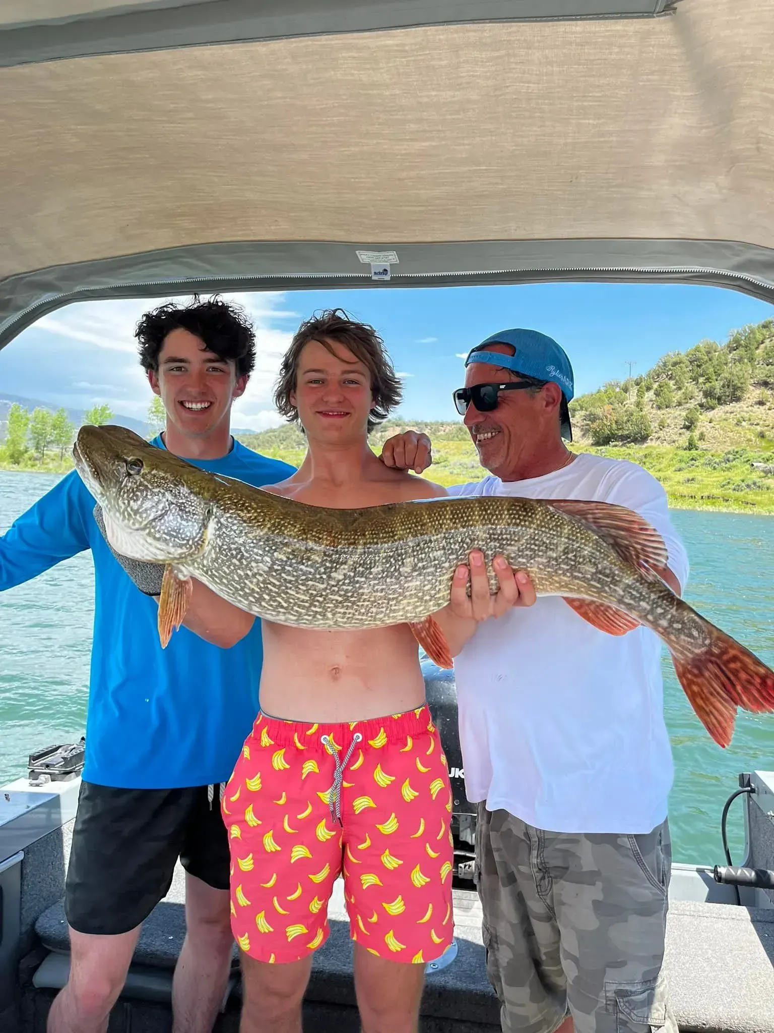 Three people on a boat holding a large fish. They are smiling. Lake in background.