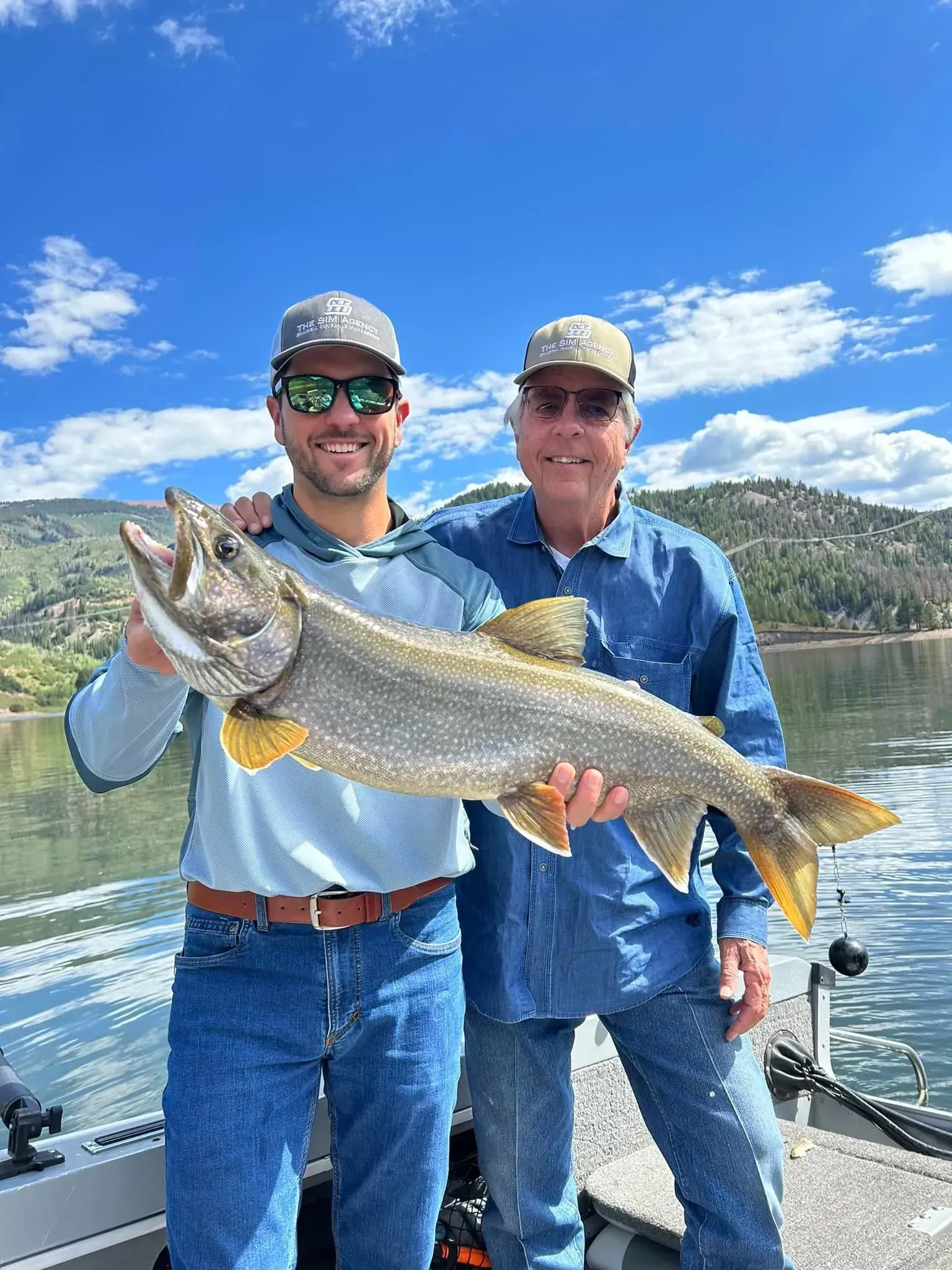 Two men on a boat hold up a large fish, lake and mountains in the background. Sunny day.