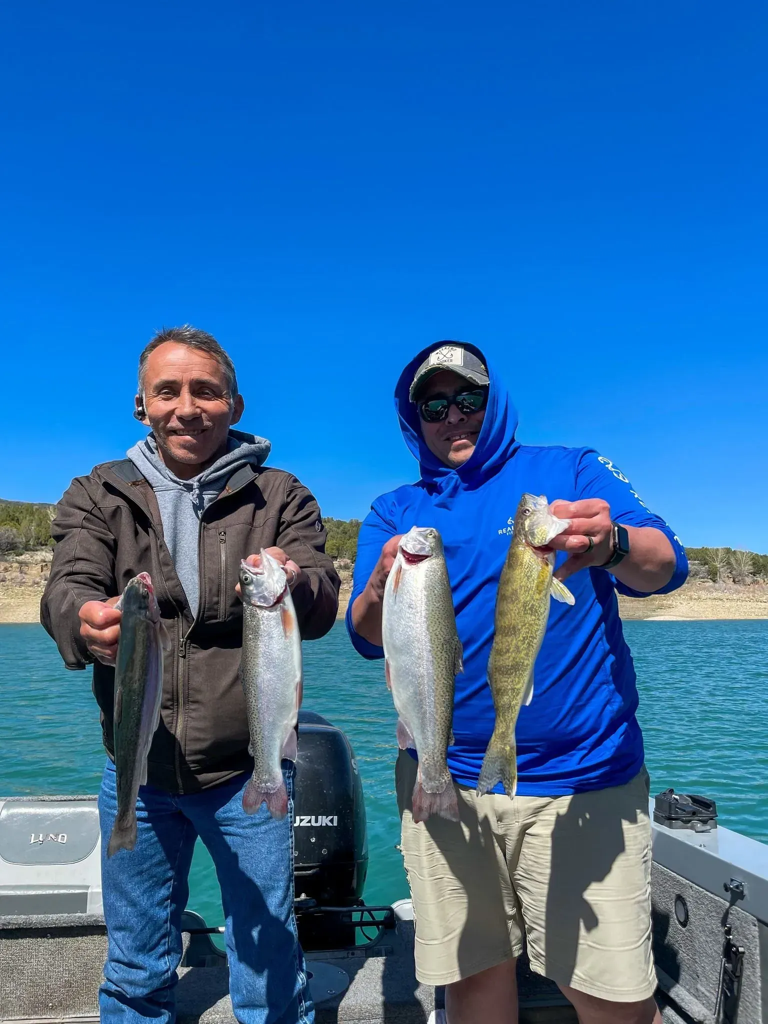 Two men on a boat, holding up freshly caught fish against a blue sky over water.