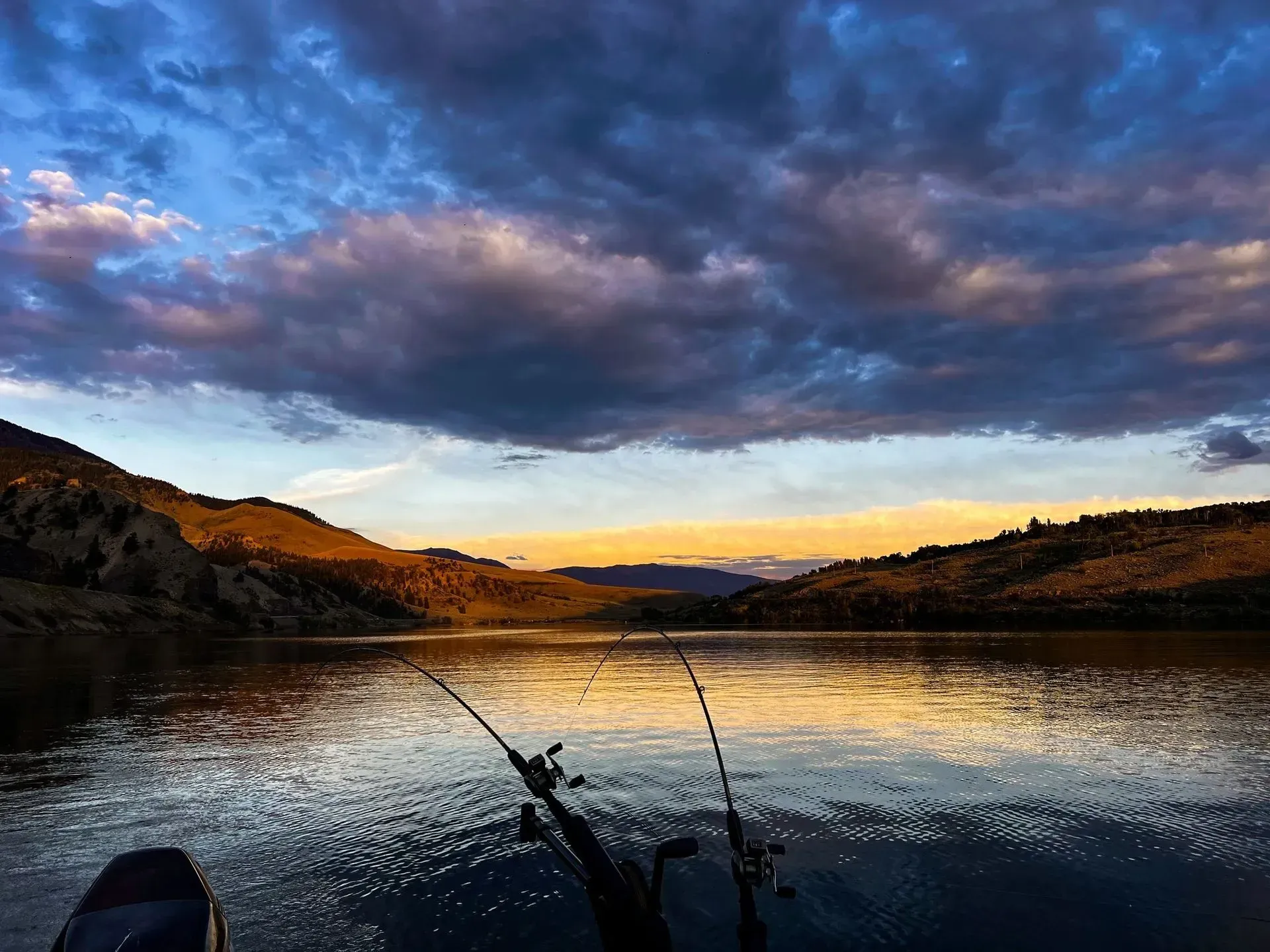 Fishing rods on a boat overlooking a calm lake at sunset. Golden light reflects on water, blue and purple clouds above hills.