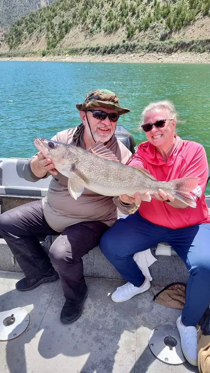 Two people holding a large fish on a boat; lake and mountains in the background.