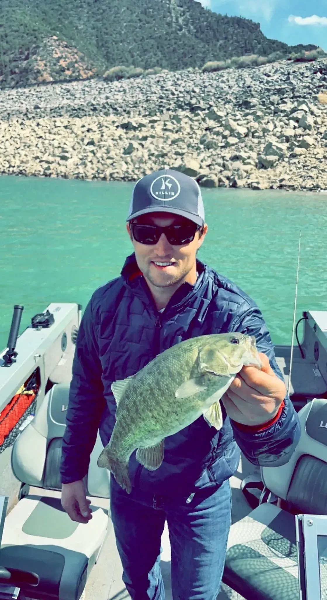 Man holding up a fish on a boat, blue water, mountains in the background.