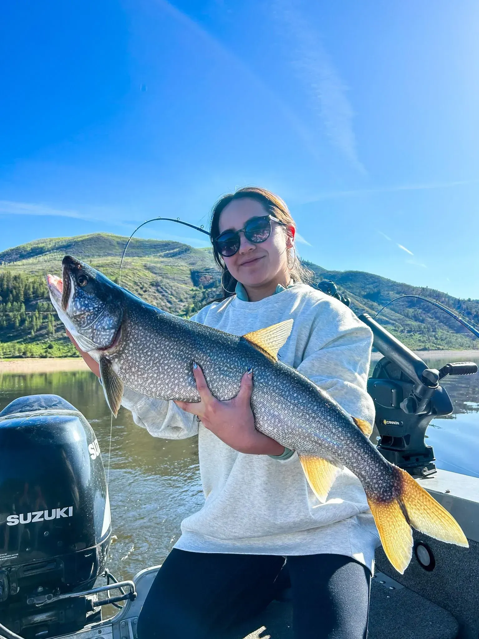 Woman on a boat holding a large fish with a yellow tail, sunny day, mountains in background.