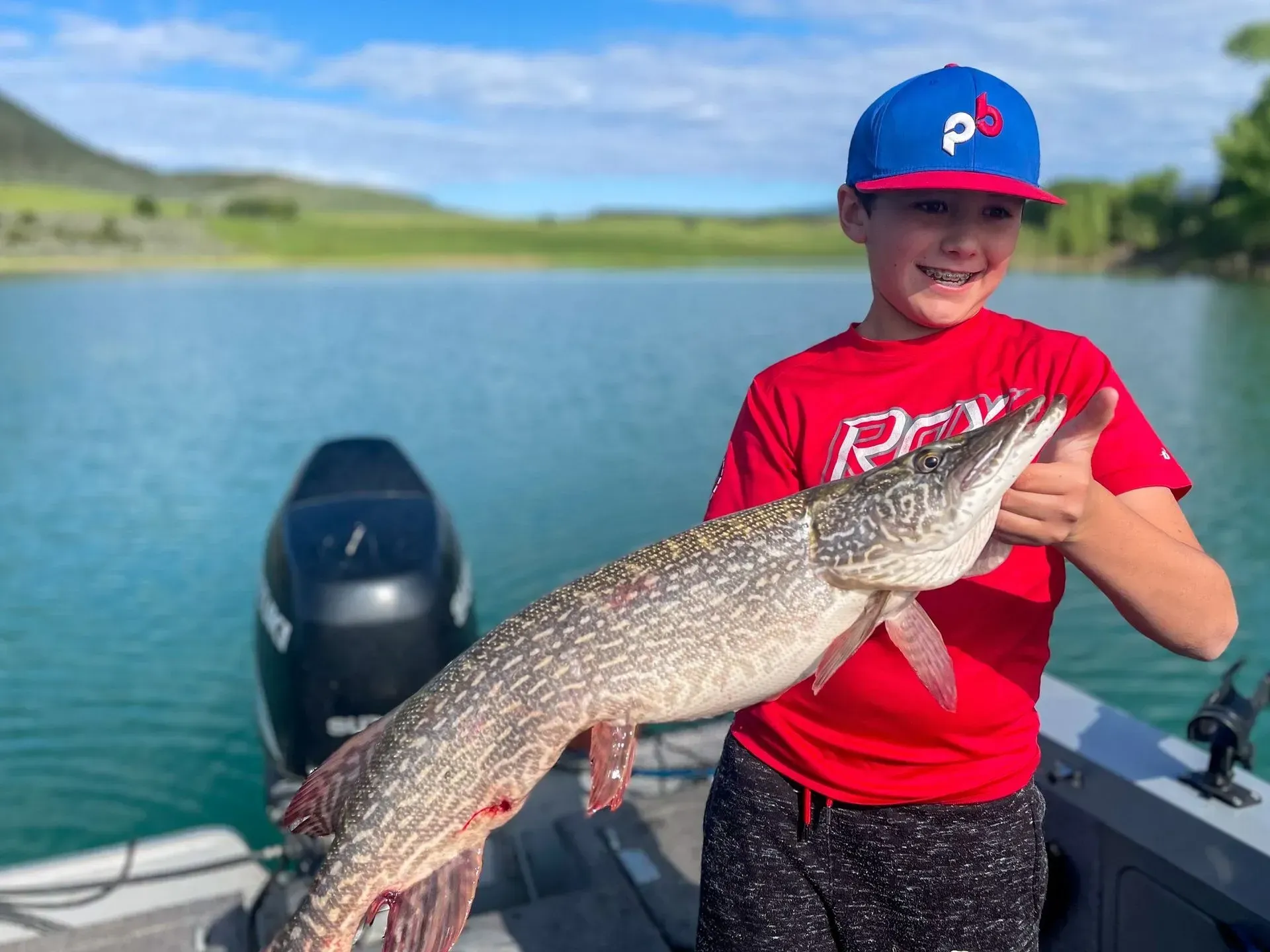 Boy on boat holds up a large fish he caught, lake in background, sunny day.
