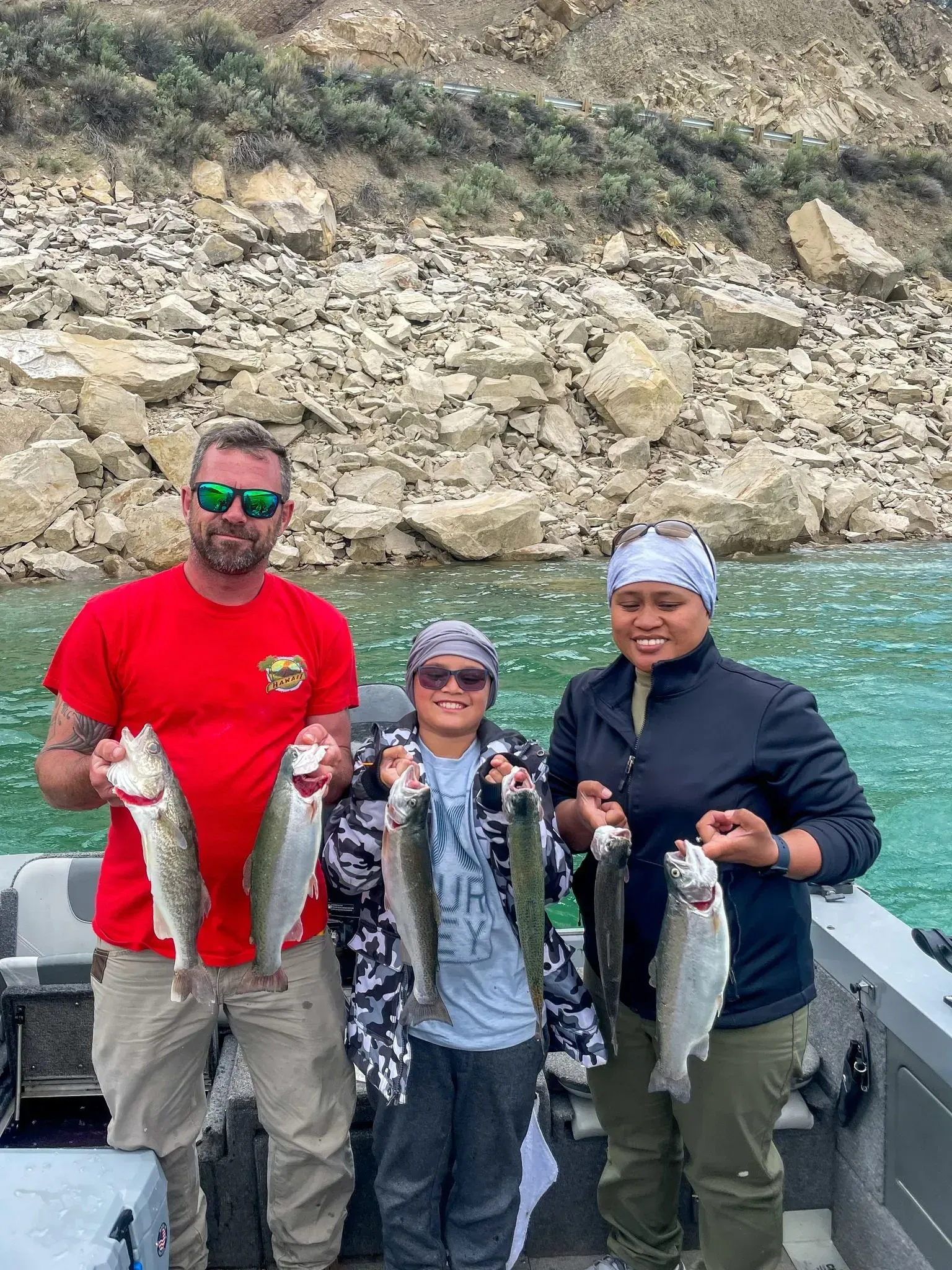 Three people on a boat holding fish they caught, with a rocky hillside and water in the background.