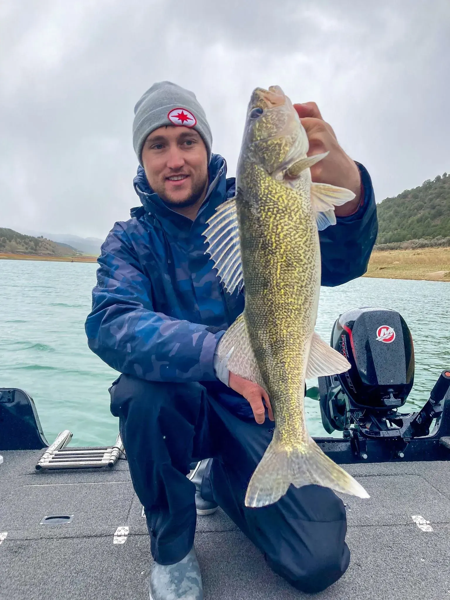 Man on a boat holds up a large walleye fish, cloudy water and distant hills in the background.