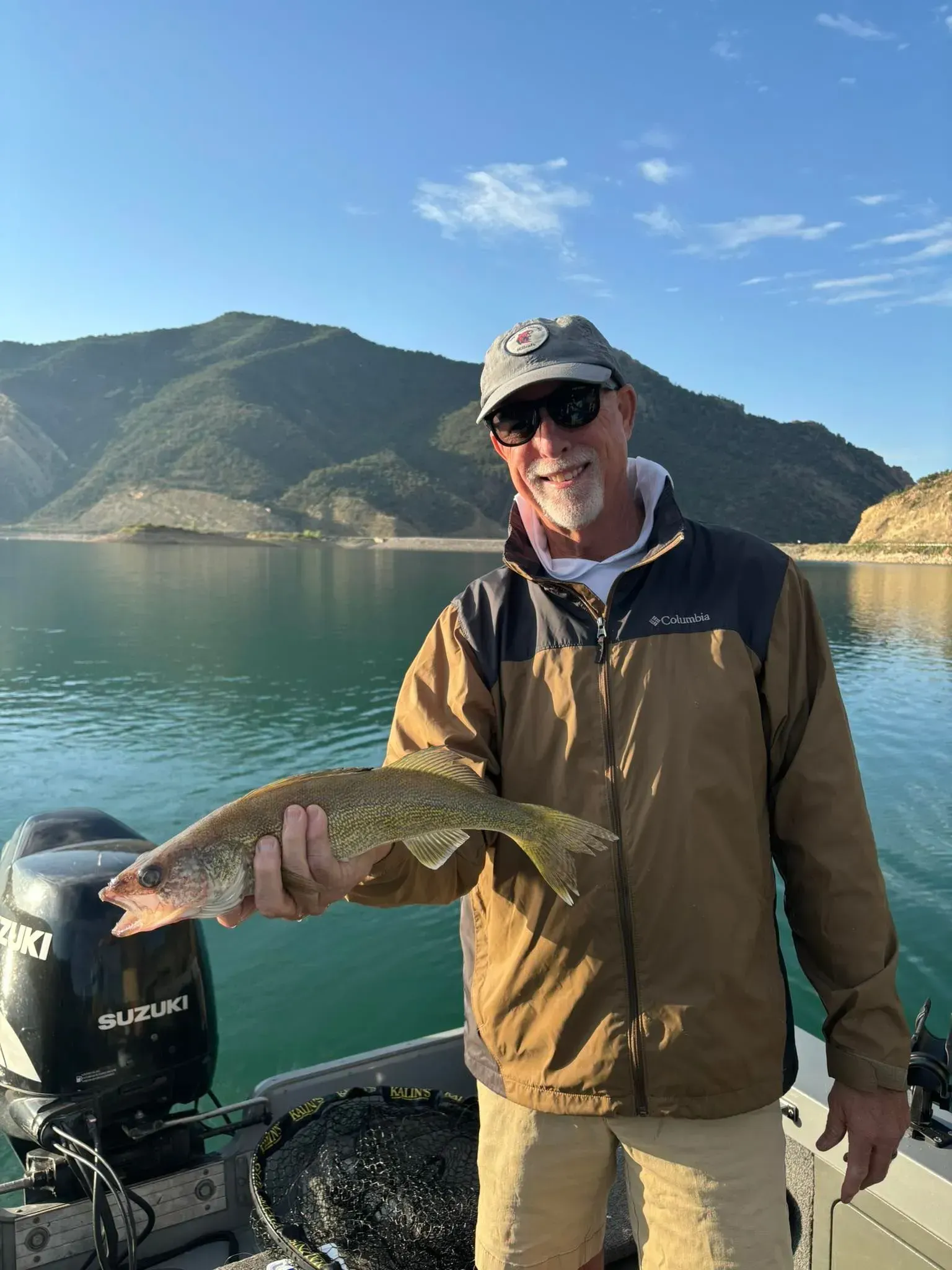 Man on boat holding fish, blue water and mountains in background.