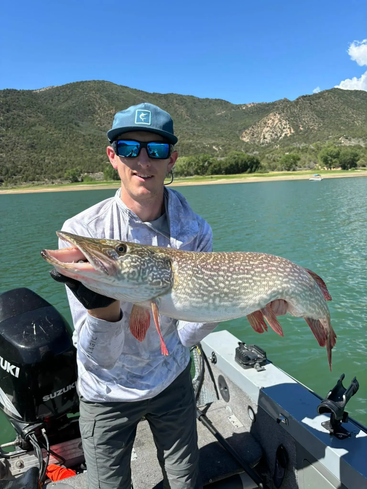 Man holding a large fish on a boat, lake and mountains visible in the background.