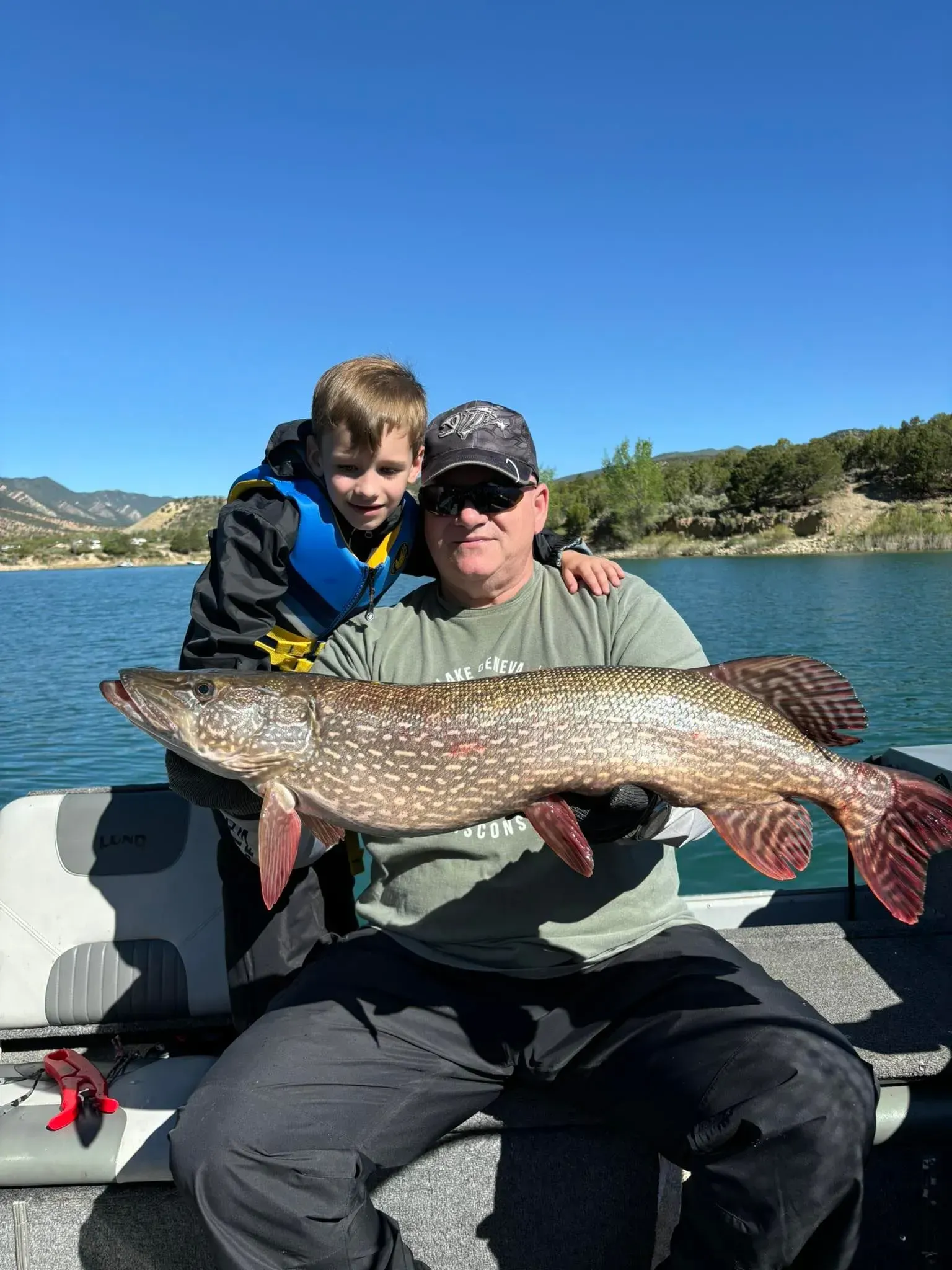 Man and boy on a boat hold a large fish. They are smiling near a lake on a sunny day.