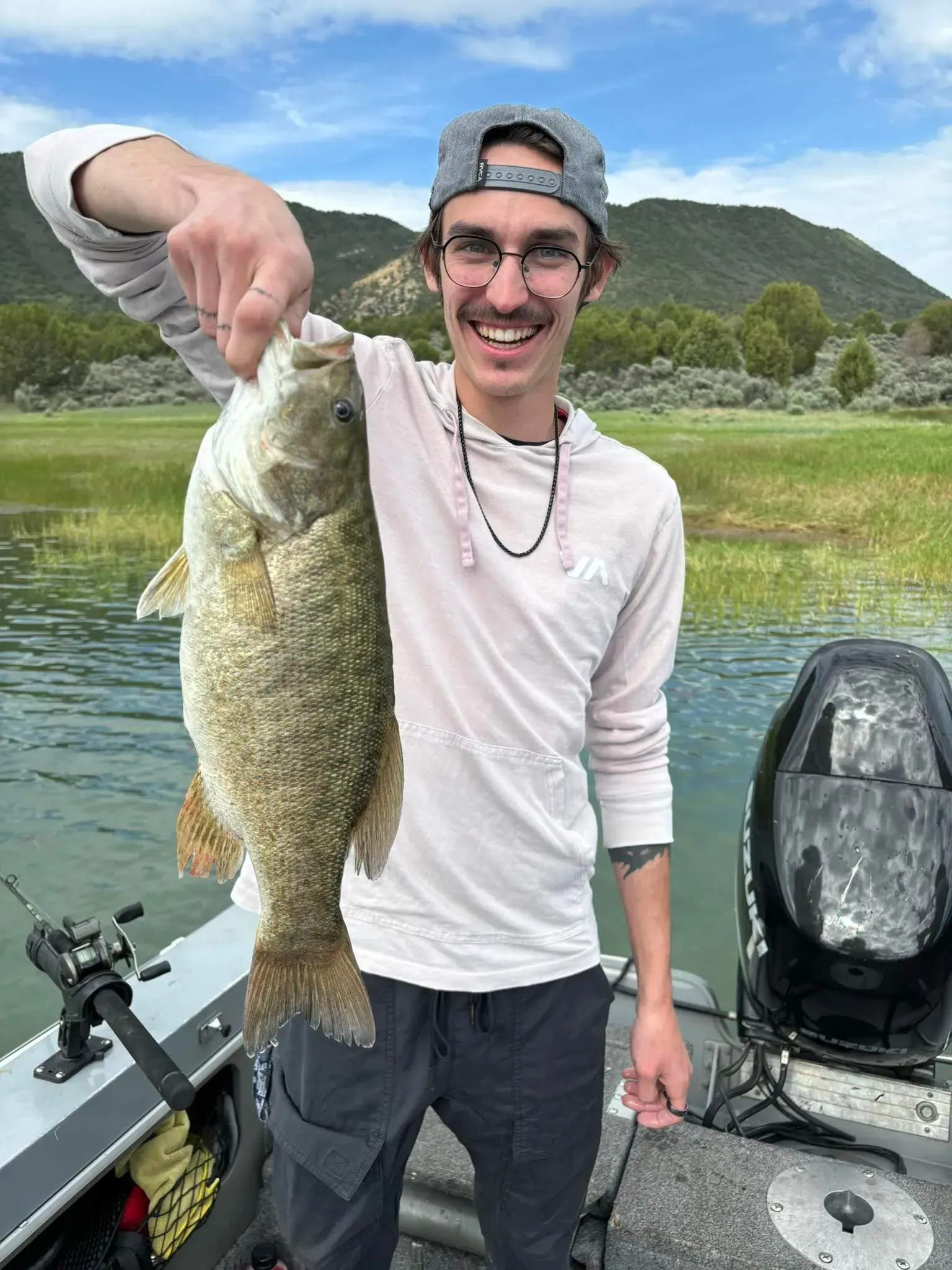 Man on boat holding up large fish, smiling. Green water, mountains in background.