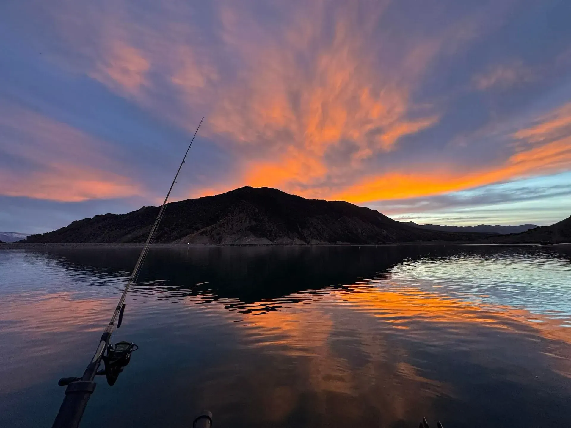 Fishing rod over calm water, reflecting a vibrant sunset of orange and blue hues. Mountain in the distance.