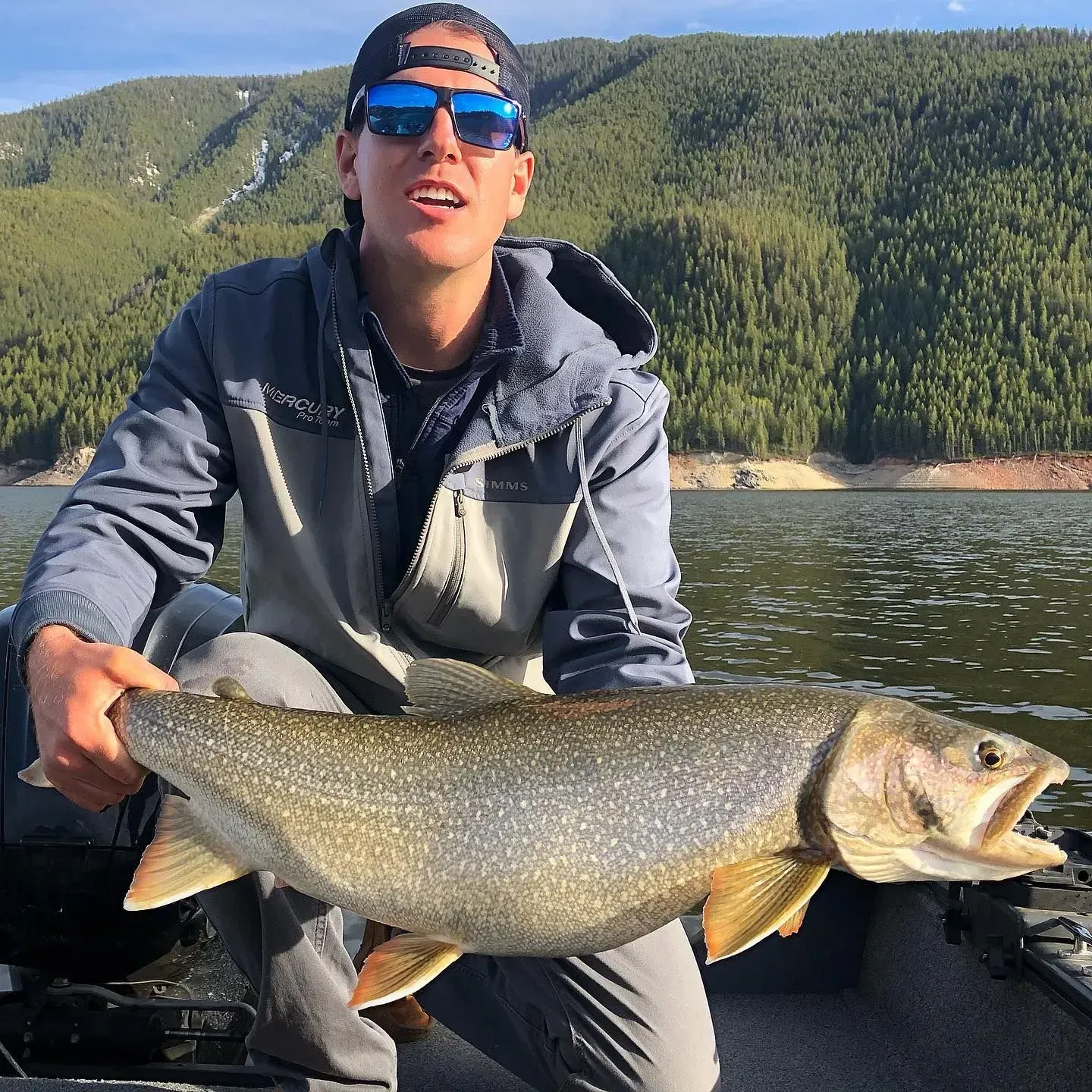 Man holding a large fish on a boat, mountains in background.
