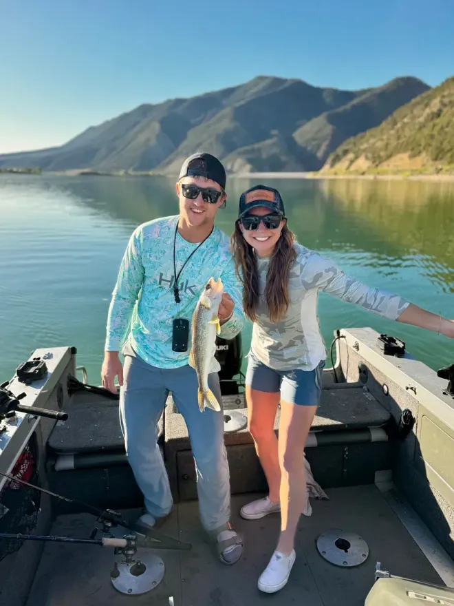 Two people holding a fish on a boat in a lake with mountains in the background.
