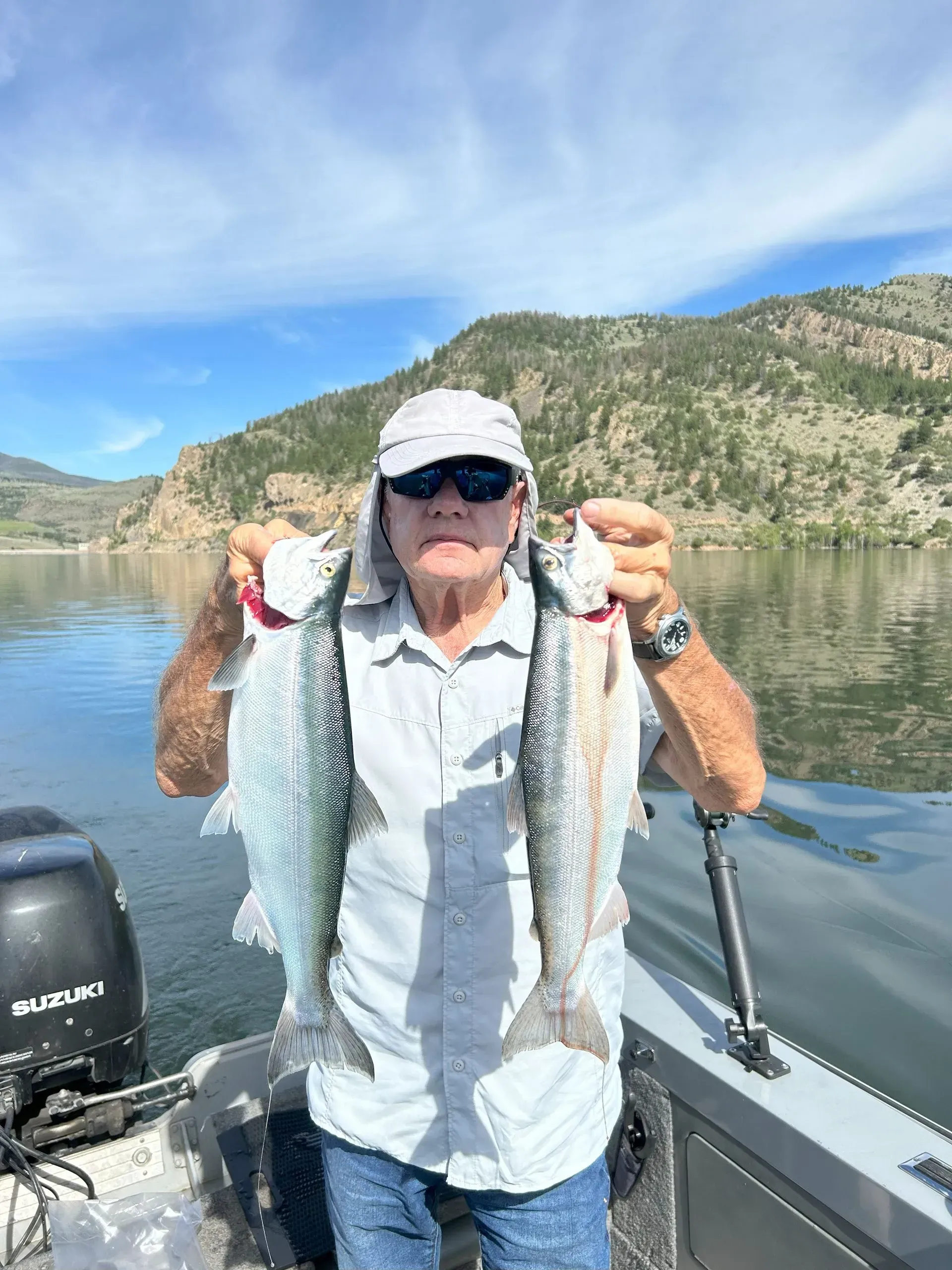 Man on a boat holds up two large fish, lake and mountains in the background. Sunny day.