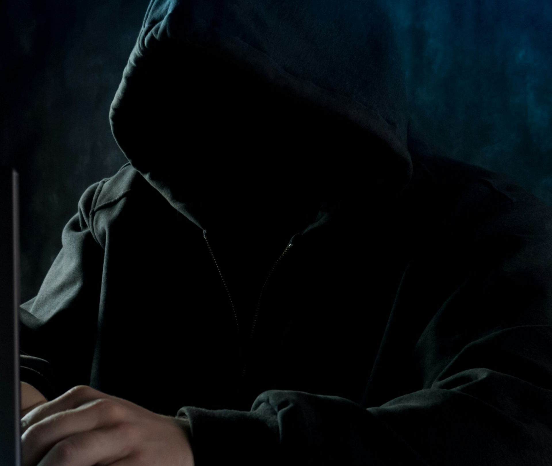 Person in a dark hooded sweatshirt using a laptop, face obscured by shadow.