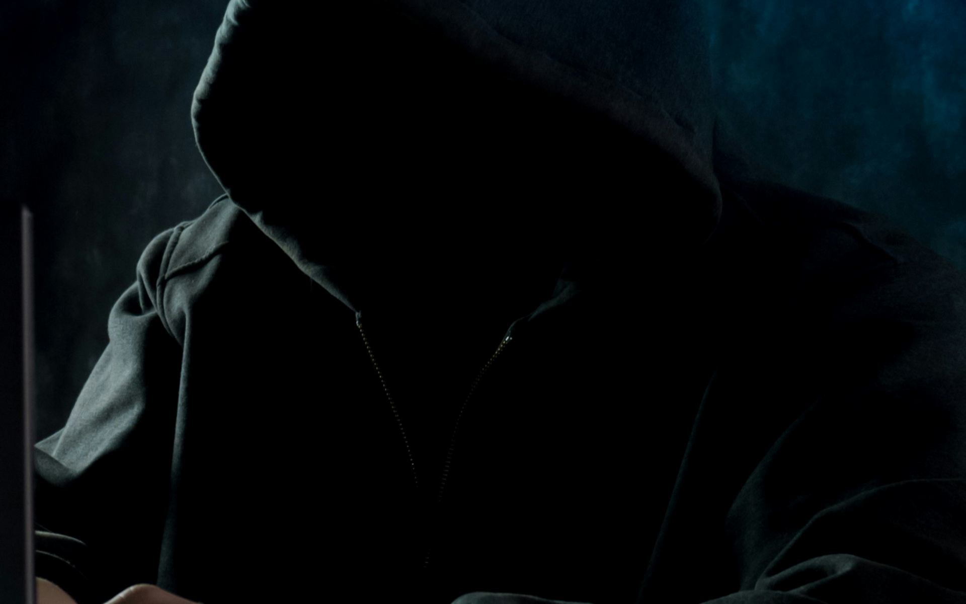 Person in a dark hooded sweatshirt using a laptop, face obscured by shadow.