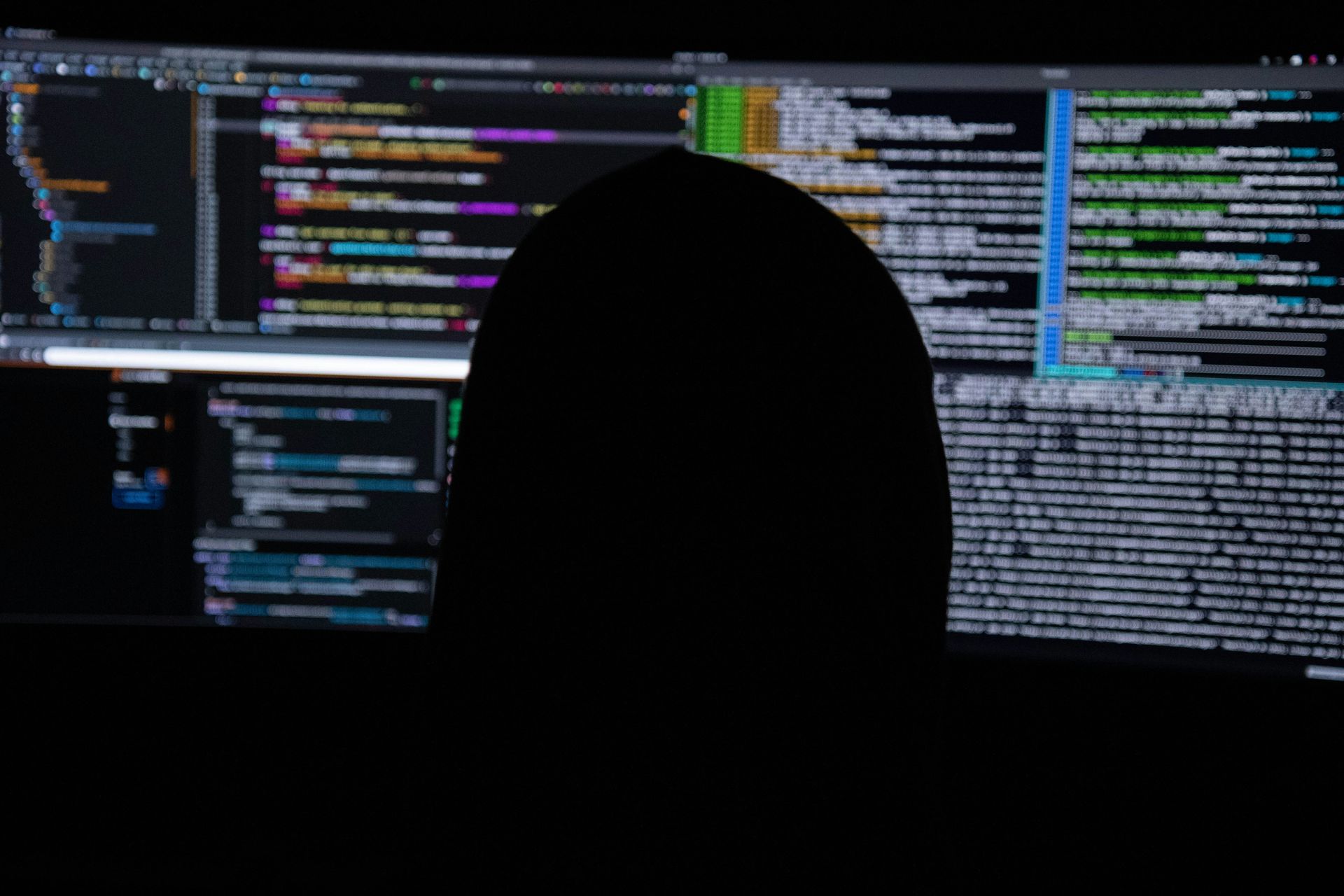 Silhouette of person in front of computer screens displaying lines of code, dark setting.