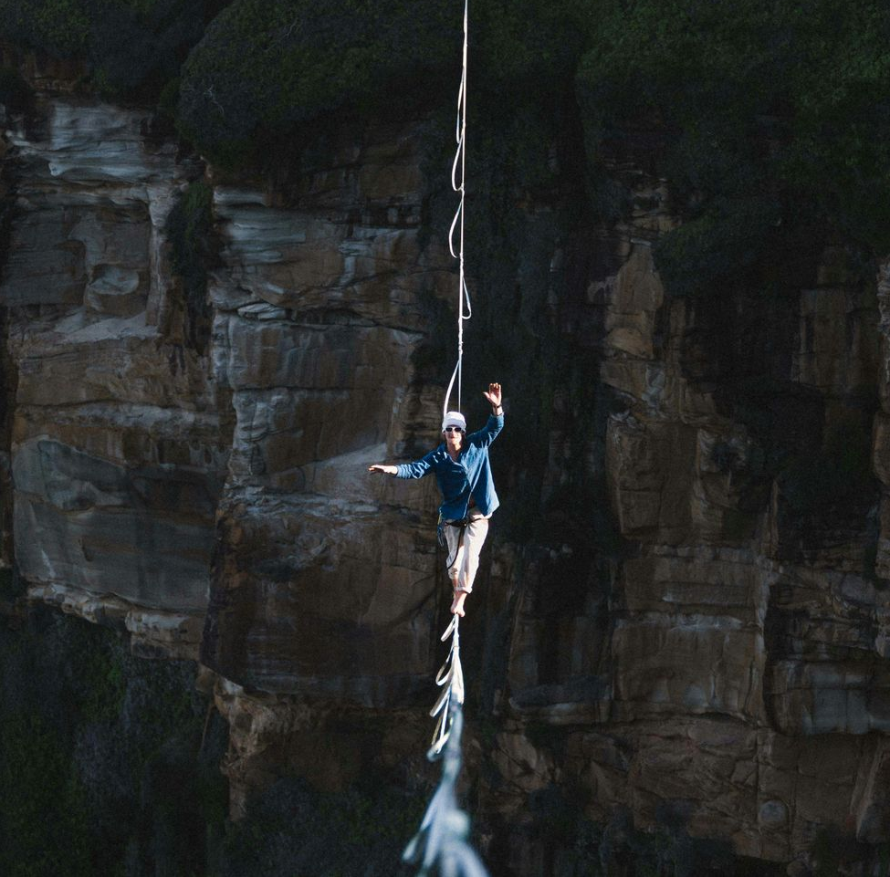 Person balances on a highline rope stretched between cliffs, arms outstretched, neutral expression.
